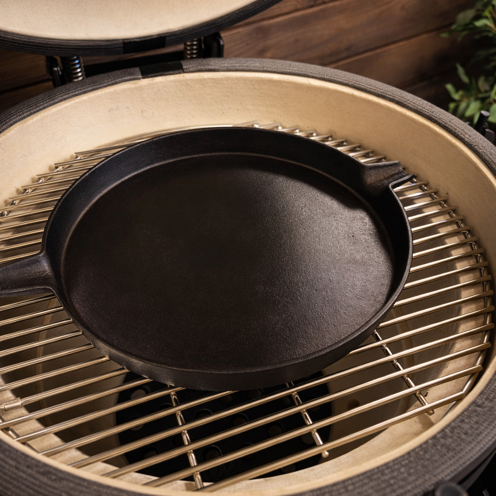 A cast iron skillet rests on a grill grate inside a ceramic cooker.