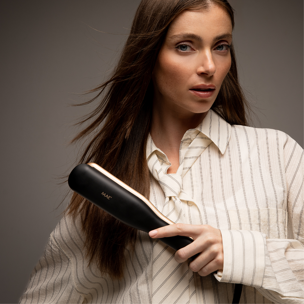 A woman with long brown hair holds a black hair straightener with gold accents.