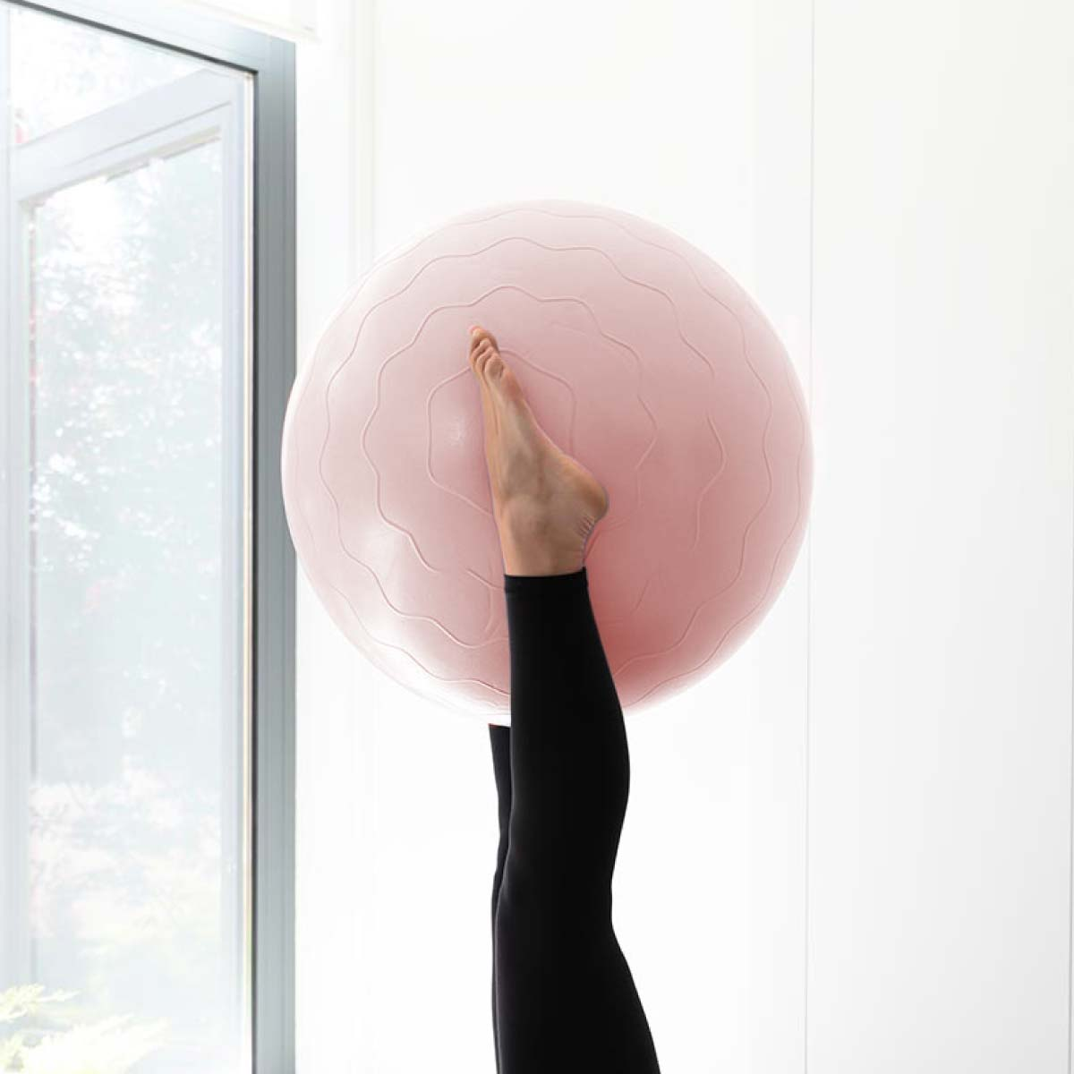 Piernas de negro con una pelota de ejercicio rosa frente a una ventana.