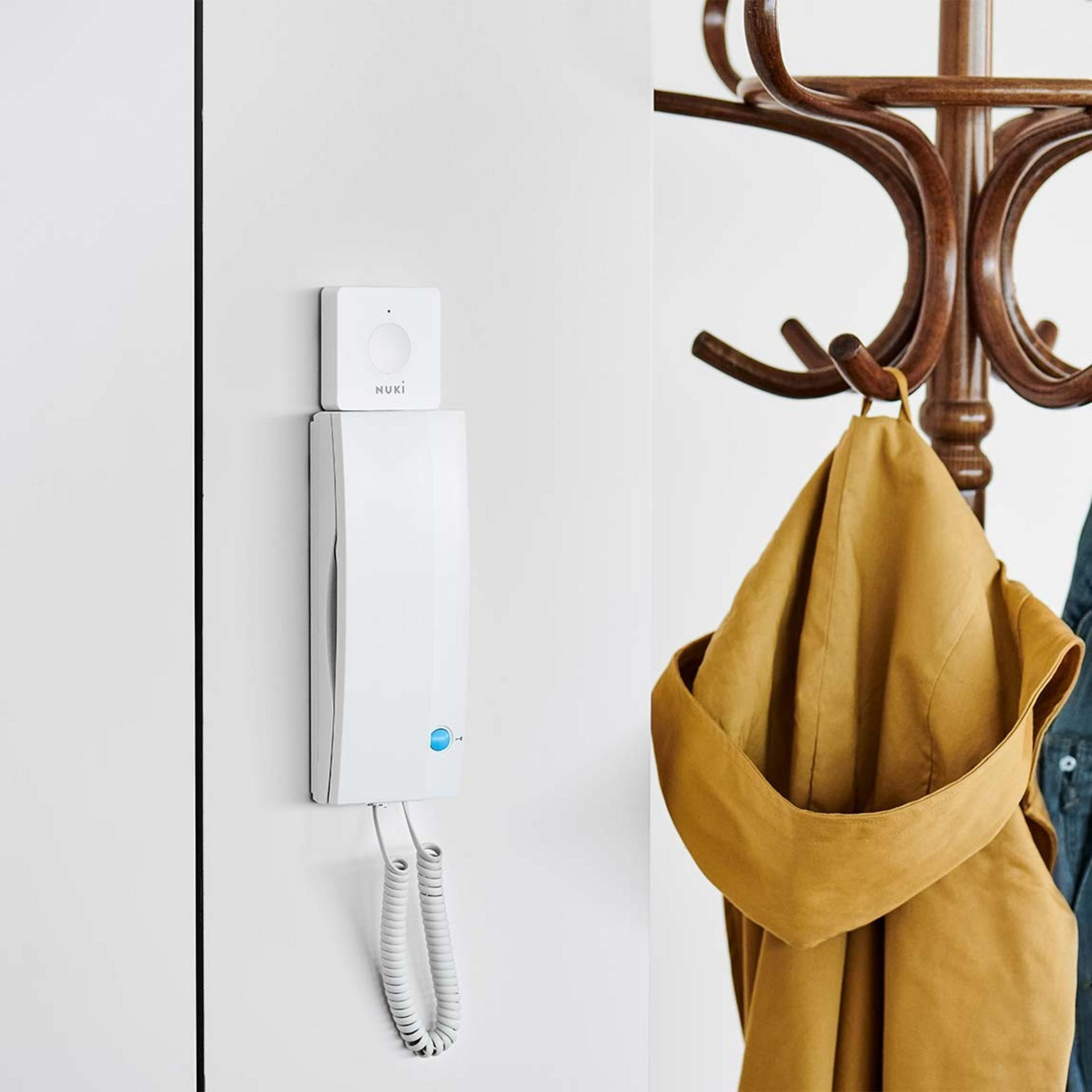 Un module d'interphone Nuki blanc sur un mur, à côté d'un porte-manteau avec des vestes.