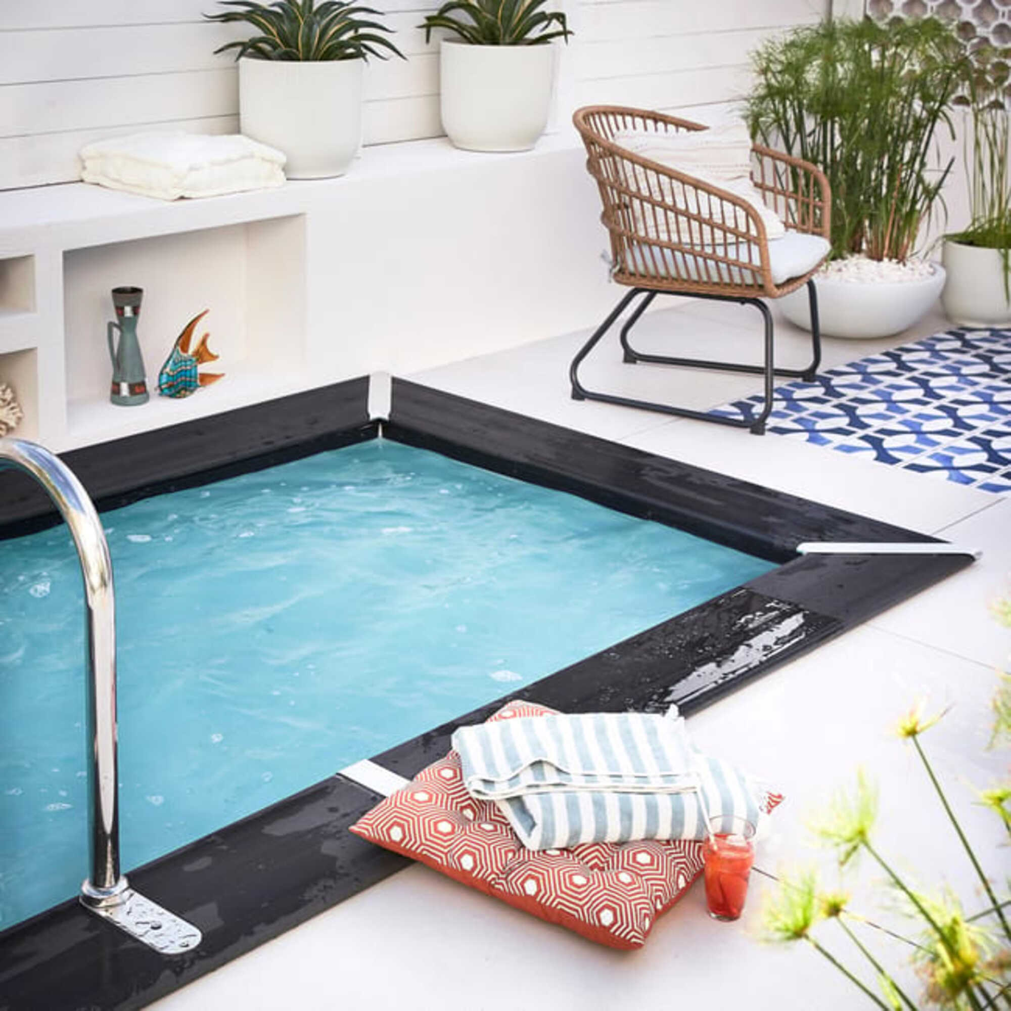 Una pequeña piscina azul con escalera cromada, junto a una silla, cojines y una bebida.