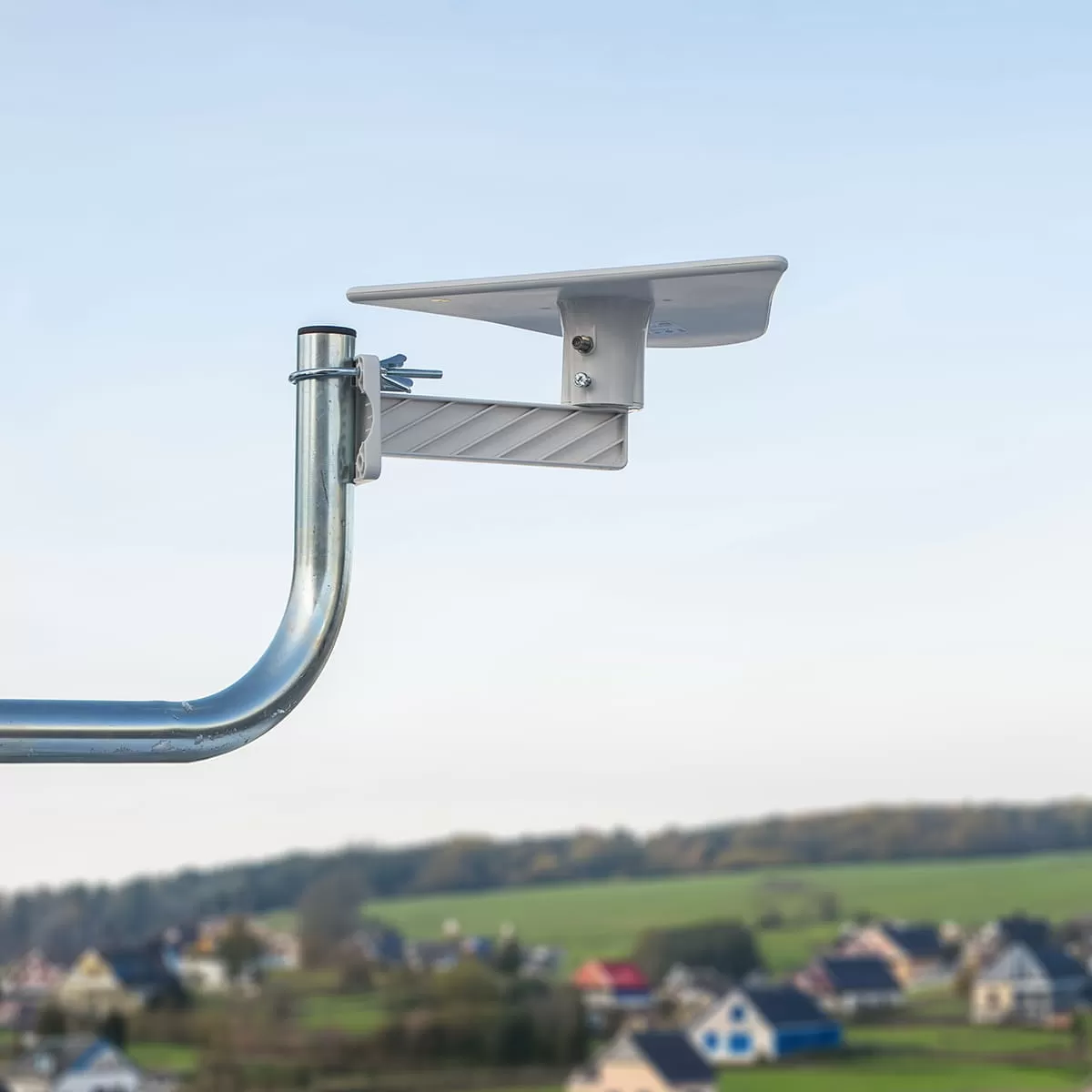 Biały uchwyt LNB satelitarnego na metalowym maszcie, nad wiejskim krajobrazem.