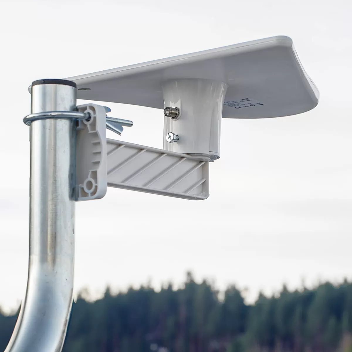 Biała antena satelitarna zamontowana na srebrnym maszcie, z sylwetką lasu w tle.