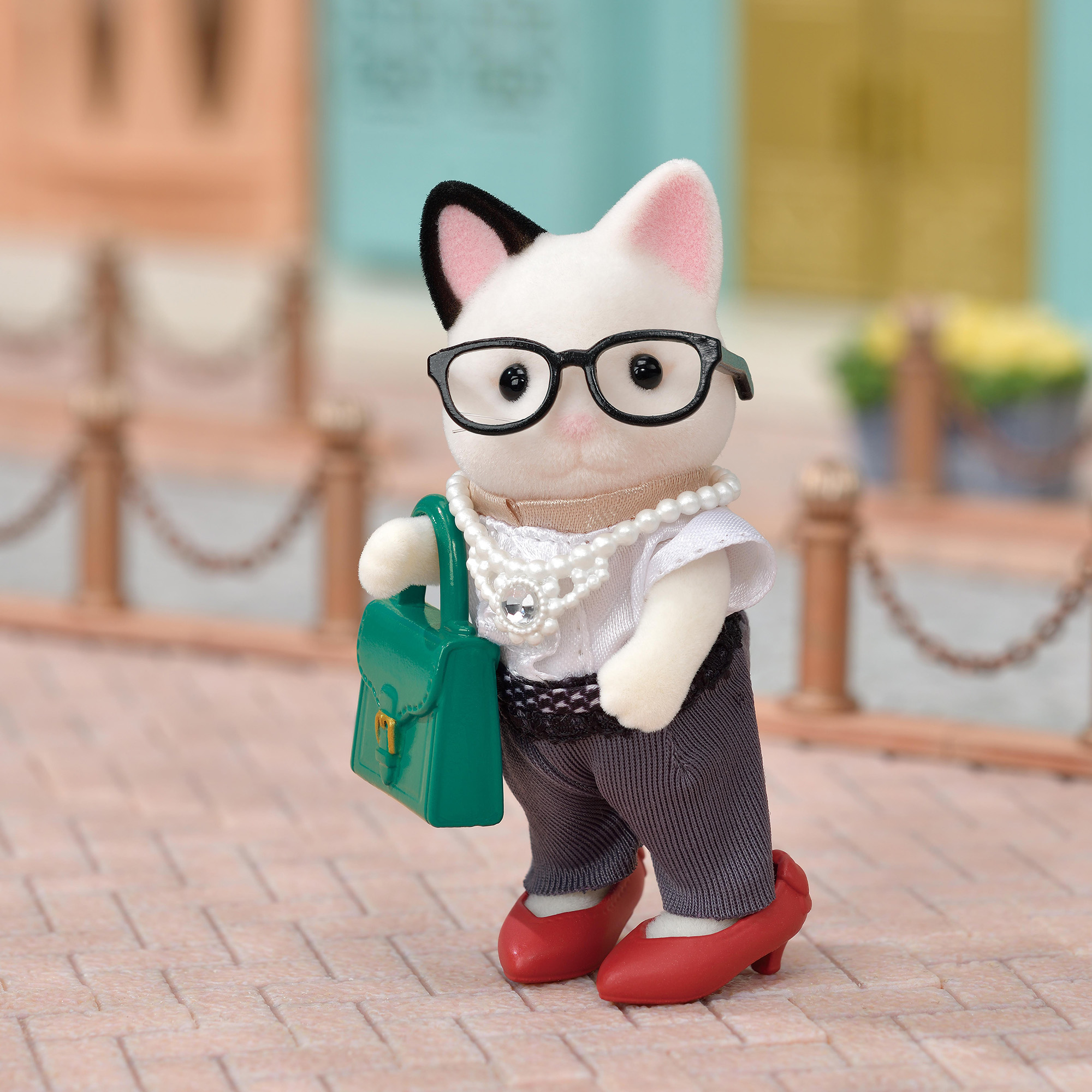Eine Katzenfigur trägt Brille, Perlen, weißes Hemd, graue Hose und rote Schuhe, hält eine grüne Tasche.