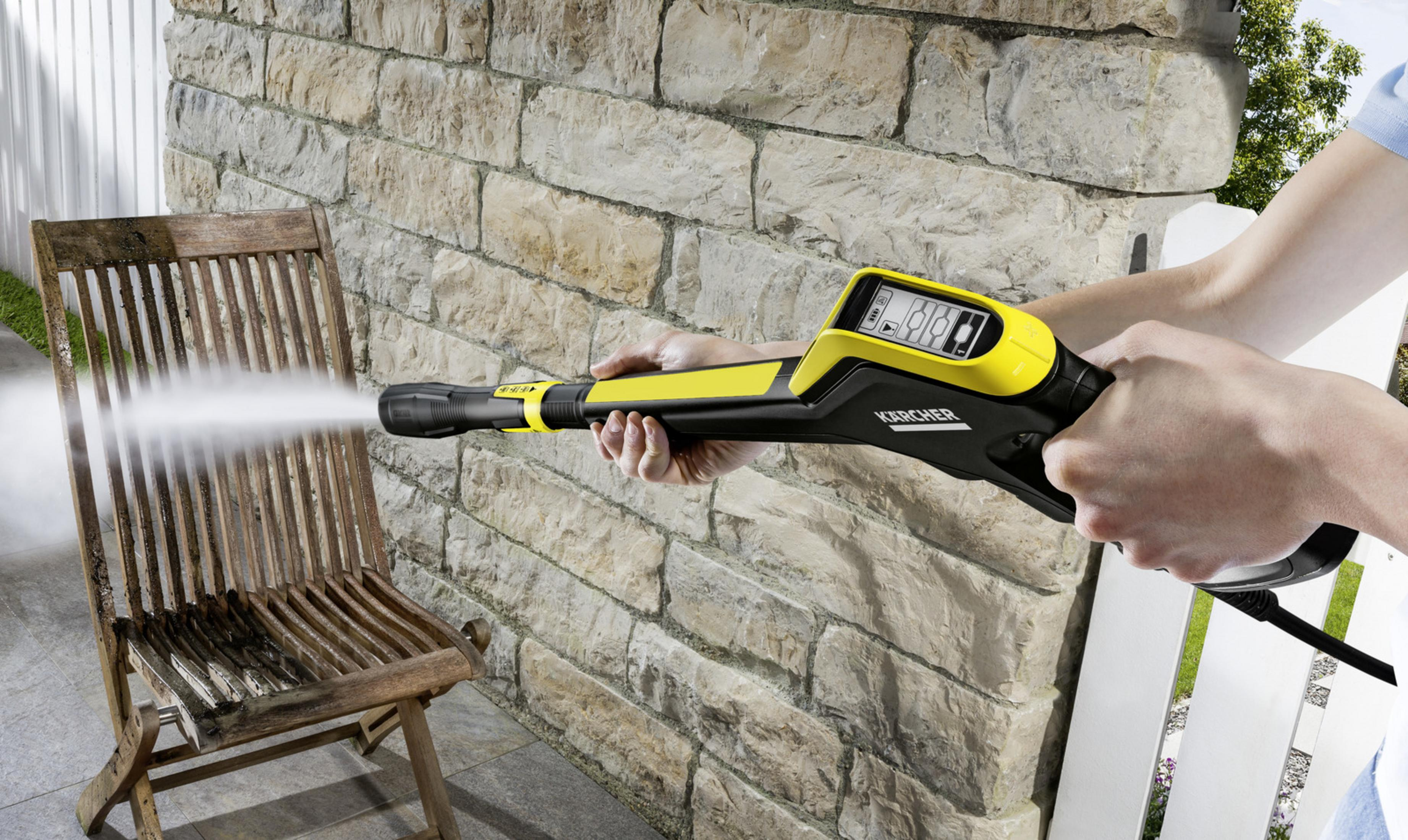 Persona pulisce sedia in legno con idropulitrice davanti a muro di pietra.