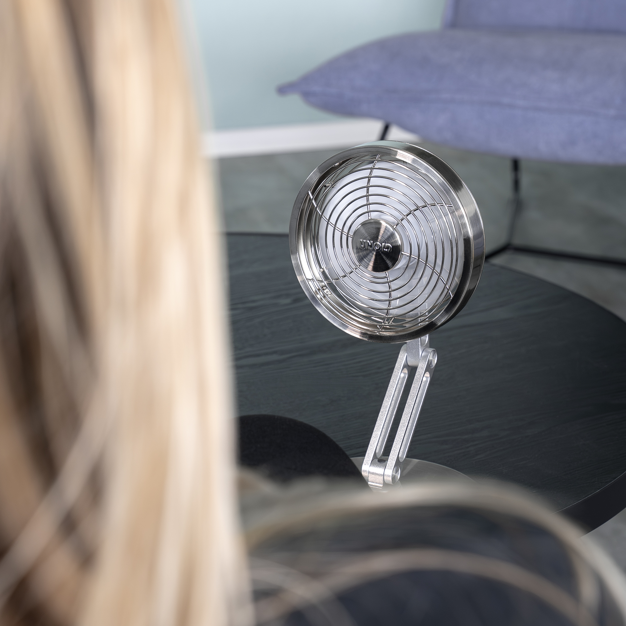 Un ventilatore da scrivania argentato è su un tavolo nero davanti a un divano blu.