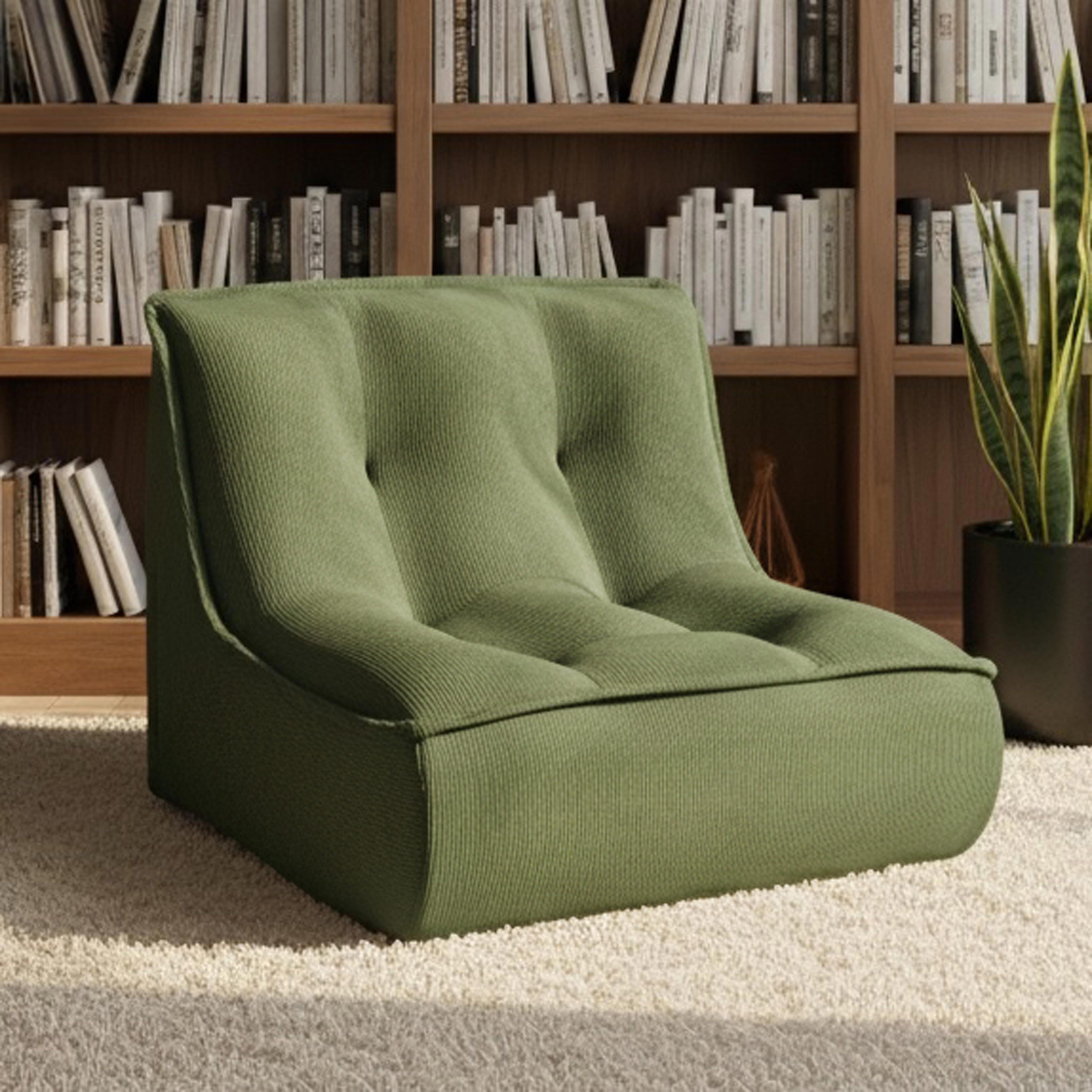 Ein grüner Cord-Sessel steht auf einem beigen Teppich vor einem Bücherregal.