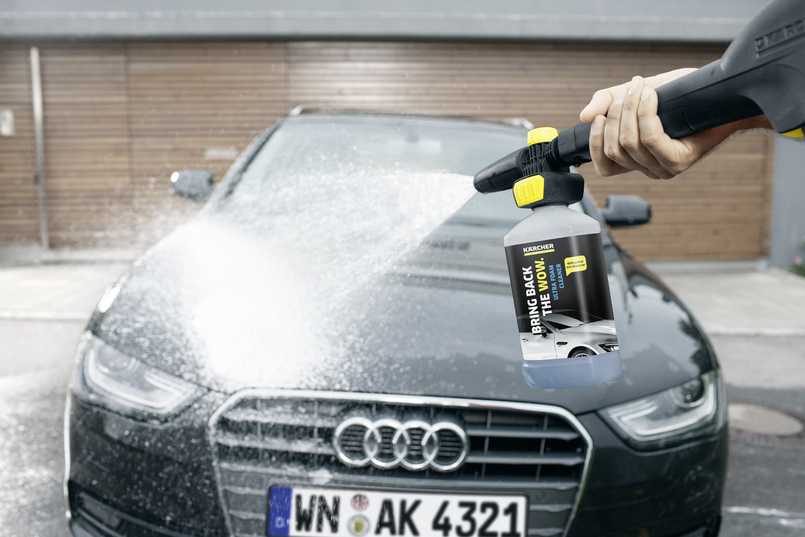 Una mano spruzza detergente schiumogeno da una bottiglia Karcher sulla parte anteriore di un'Audi scura.