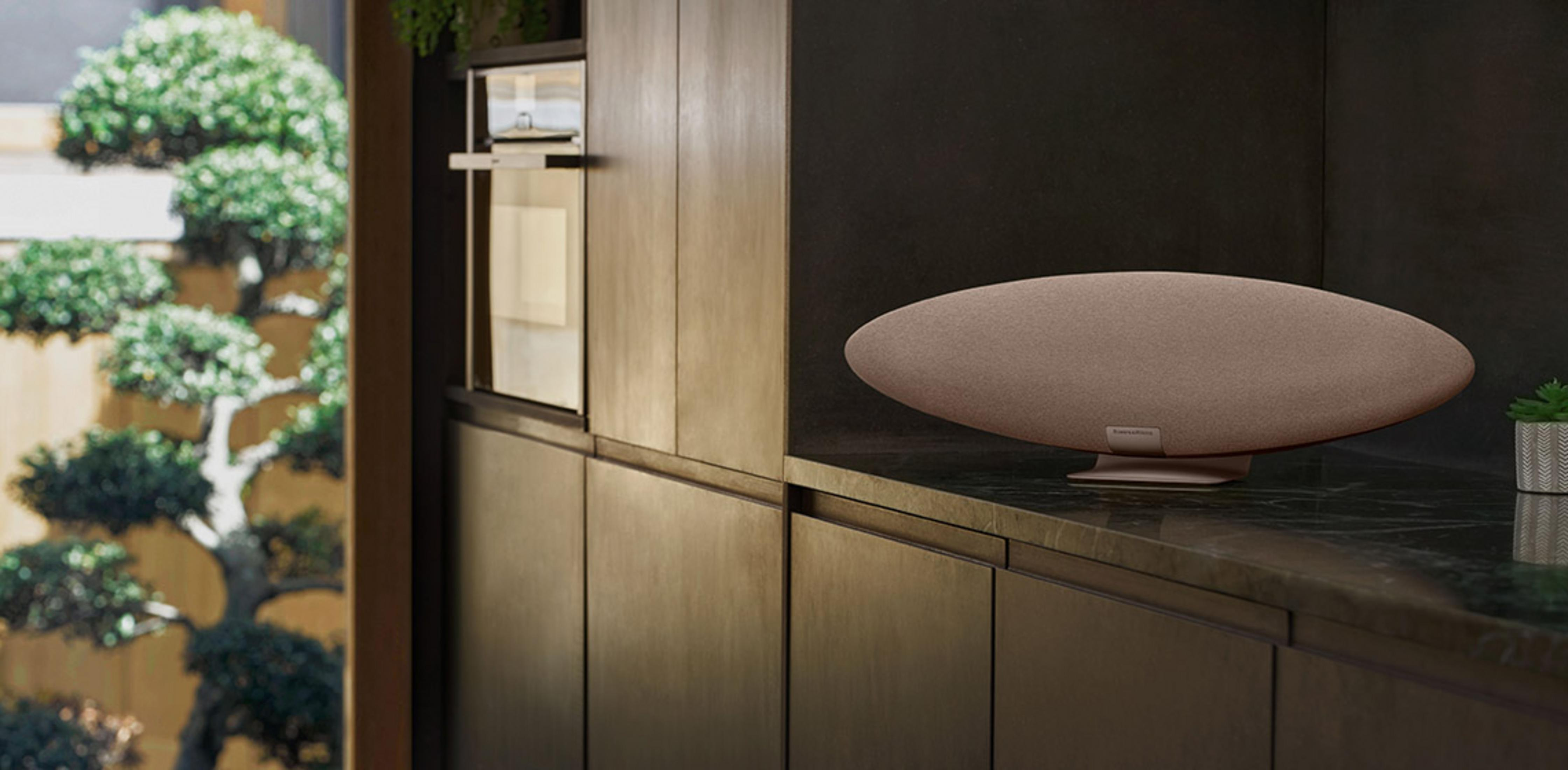 Owalny brązowy głośnik stoi na marmurowym blacie obok doniczki z małą rośliną.