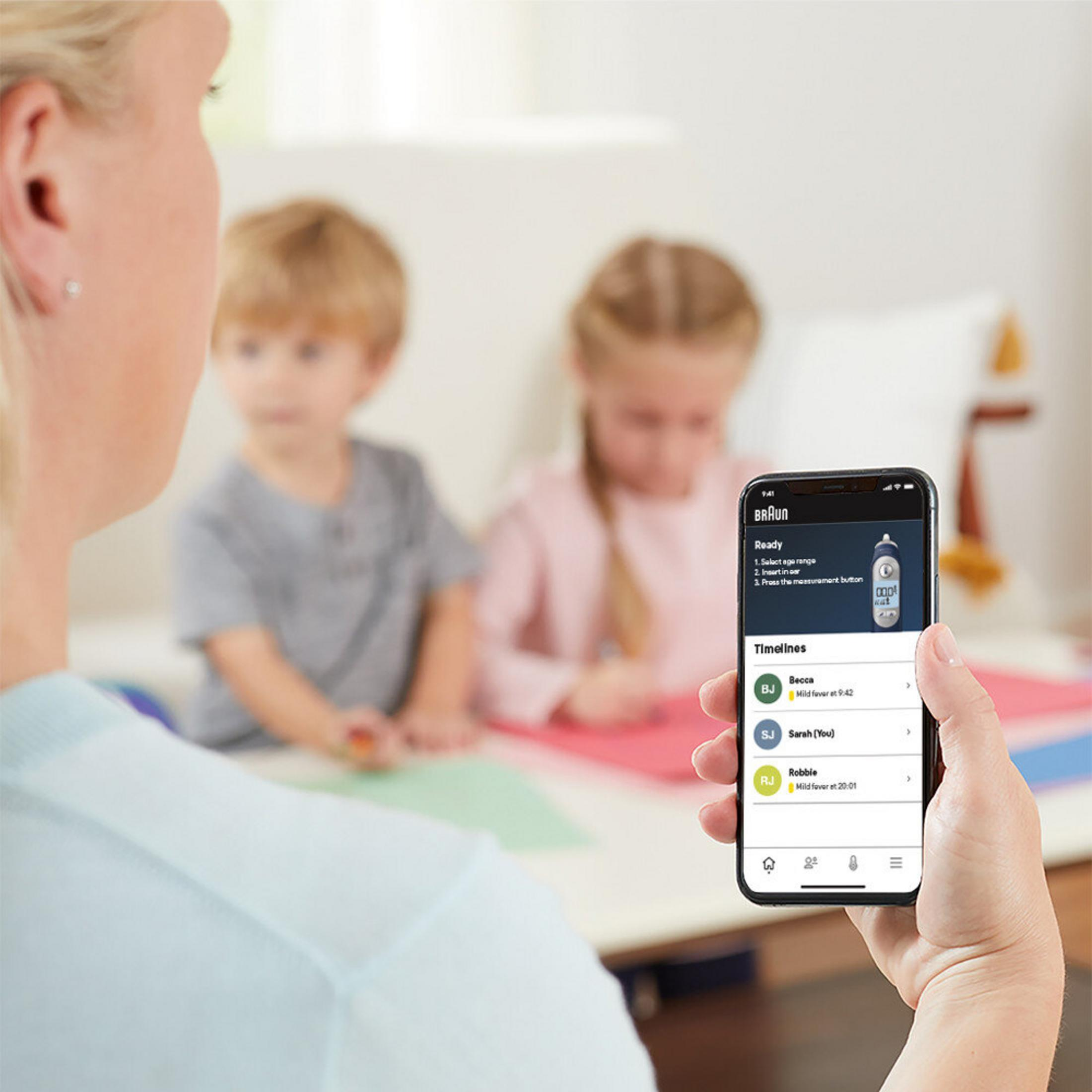 Dłoń trzyma smartfon z aplikacją BRAUN. Dwoje dzieci jest niewyraźnych w tle.