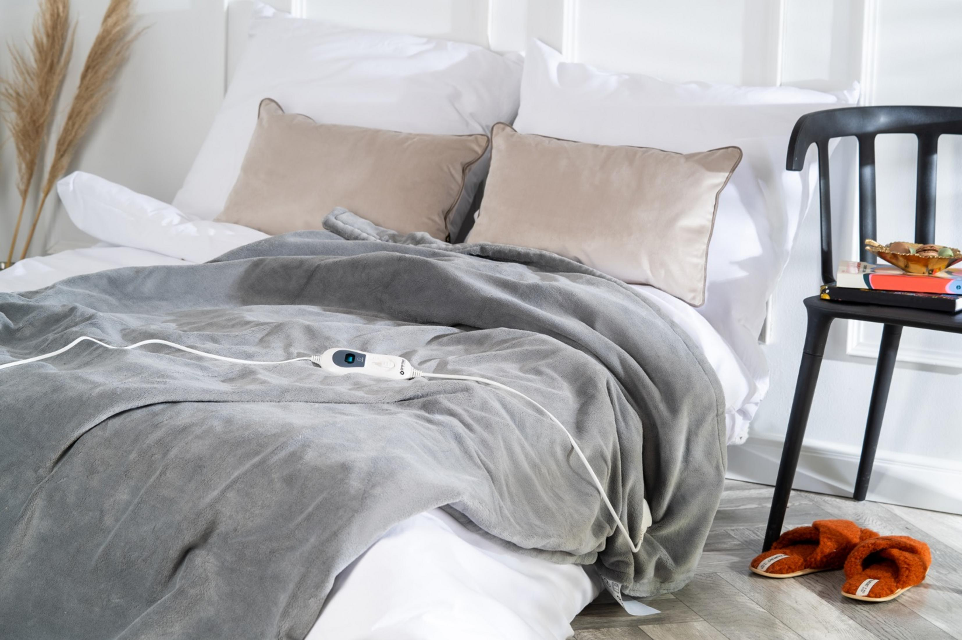 Una coperta riscaldata grigia con controller giace su un letto con cuscini. Ciabatte arancioni a terra.
