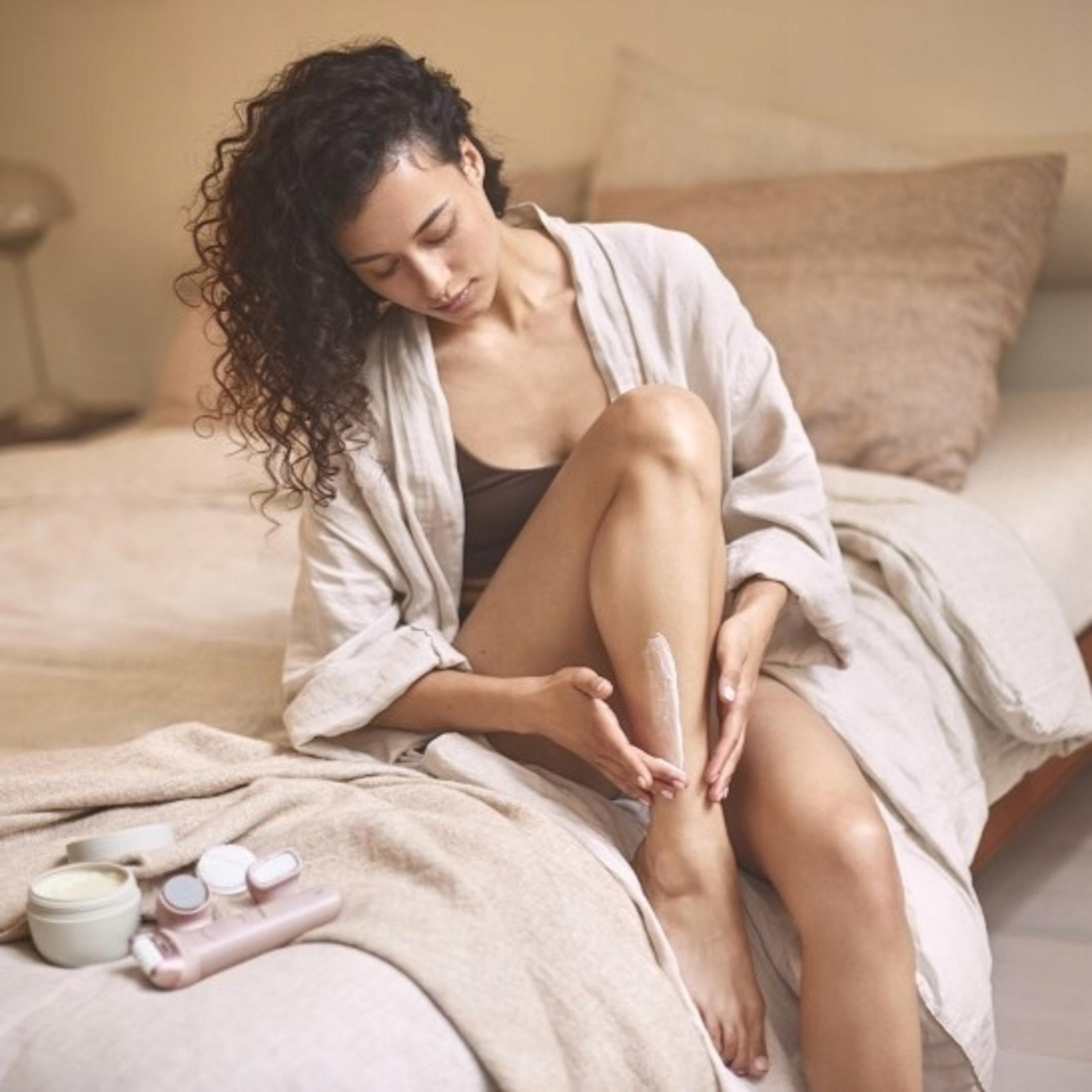 Donna con capelli ricci si applica crema sulla gamba, seduta su un letto.