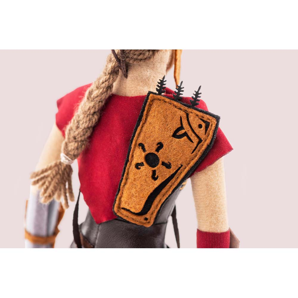 Una bambola indossa un top rosso, un gilet marrone e una treccia. Sul gilet c'è uno scudo con simboli.