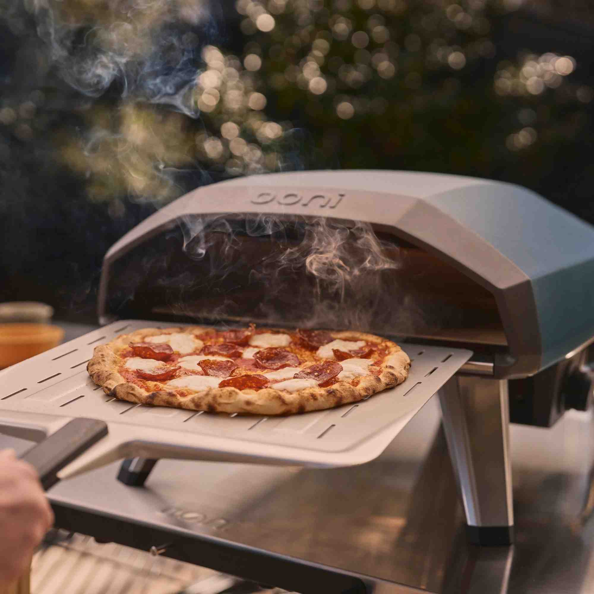 Eine Pizza mit Peperoni und Mozzarella wird aus einem Ooni-Ofen auf einem Pizzaschieber herausgenommen.