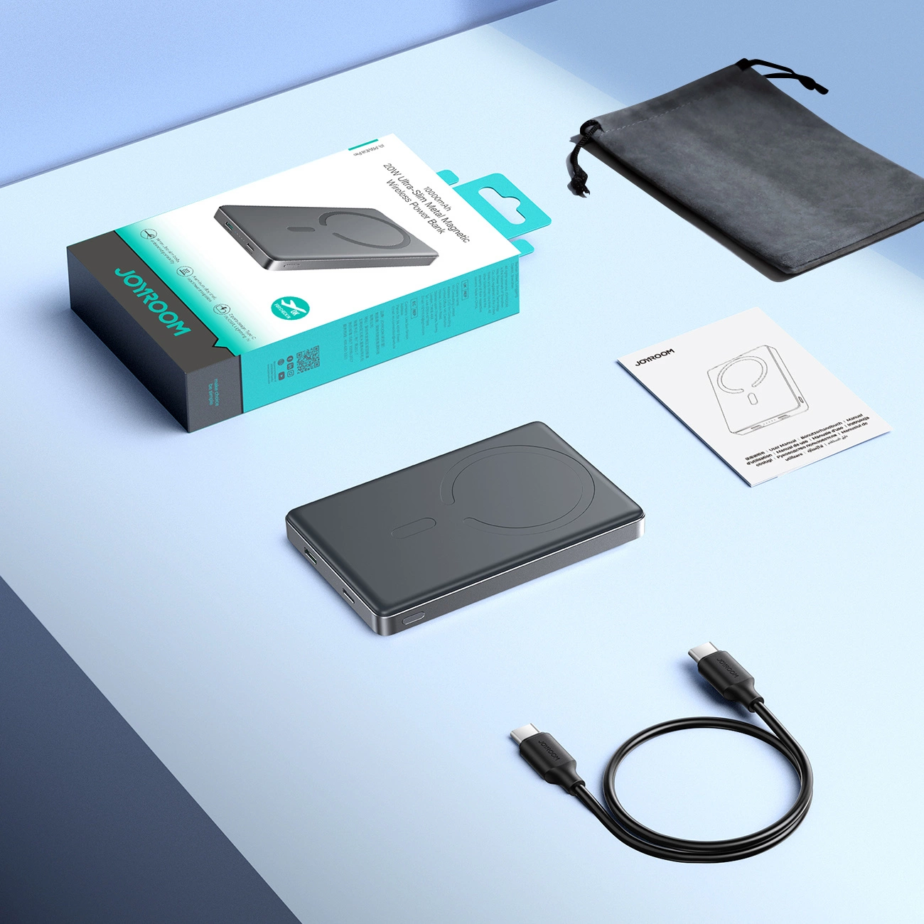 Eine graue Powerbank, ihre Verpackung, ein schwarzer Beutel, eine Bedienungsanleitung und ein Ladekabel liegen auf einer blau-grauen Oberfläche.