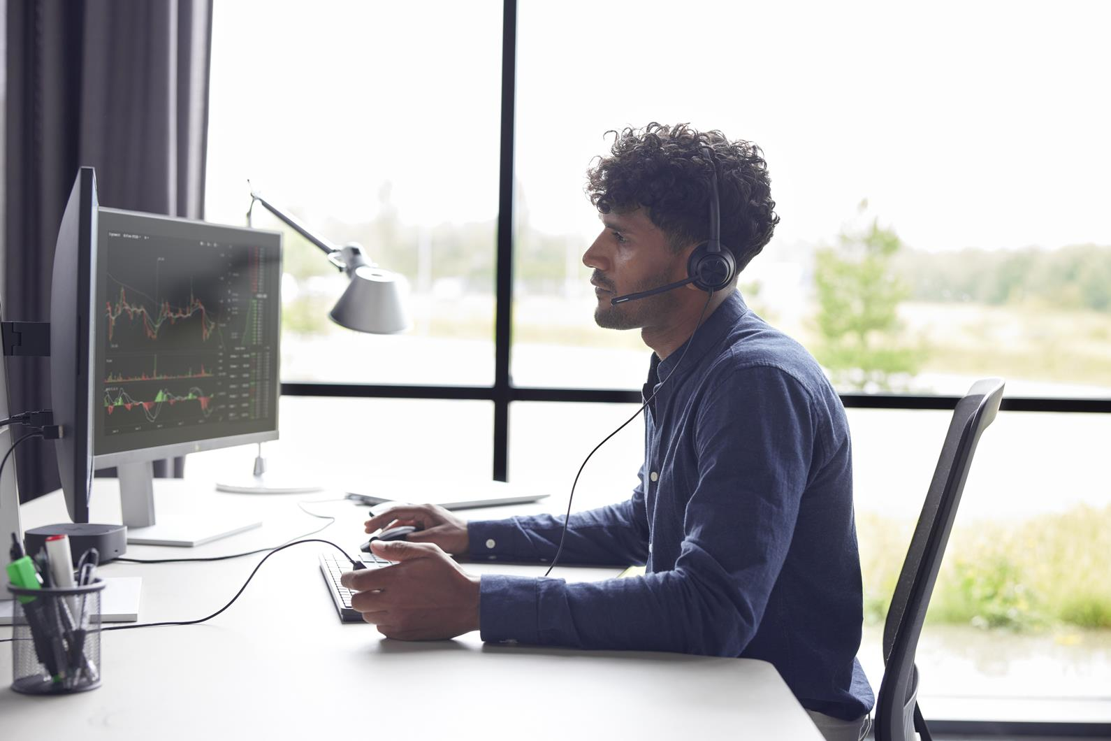Un uomo con i capelli ricci e una cuffia lavora al computer con un grafico sullo schermo, seduto vicino a una finestra.