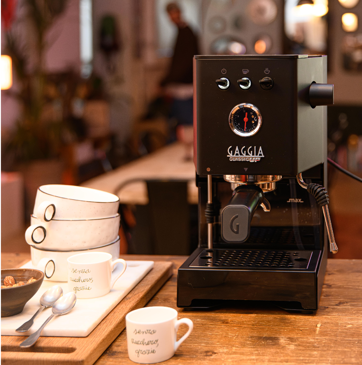 Zwarte espressomachine met kopjes en lepels op een houten tafel.