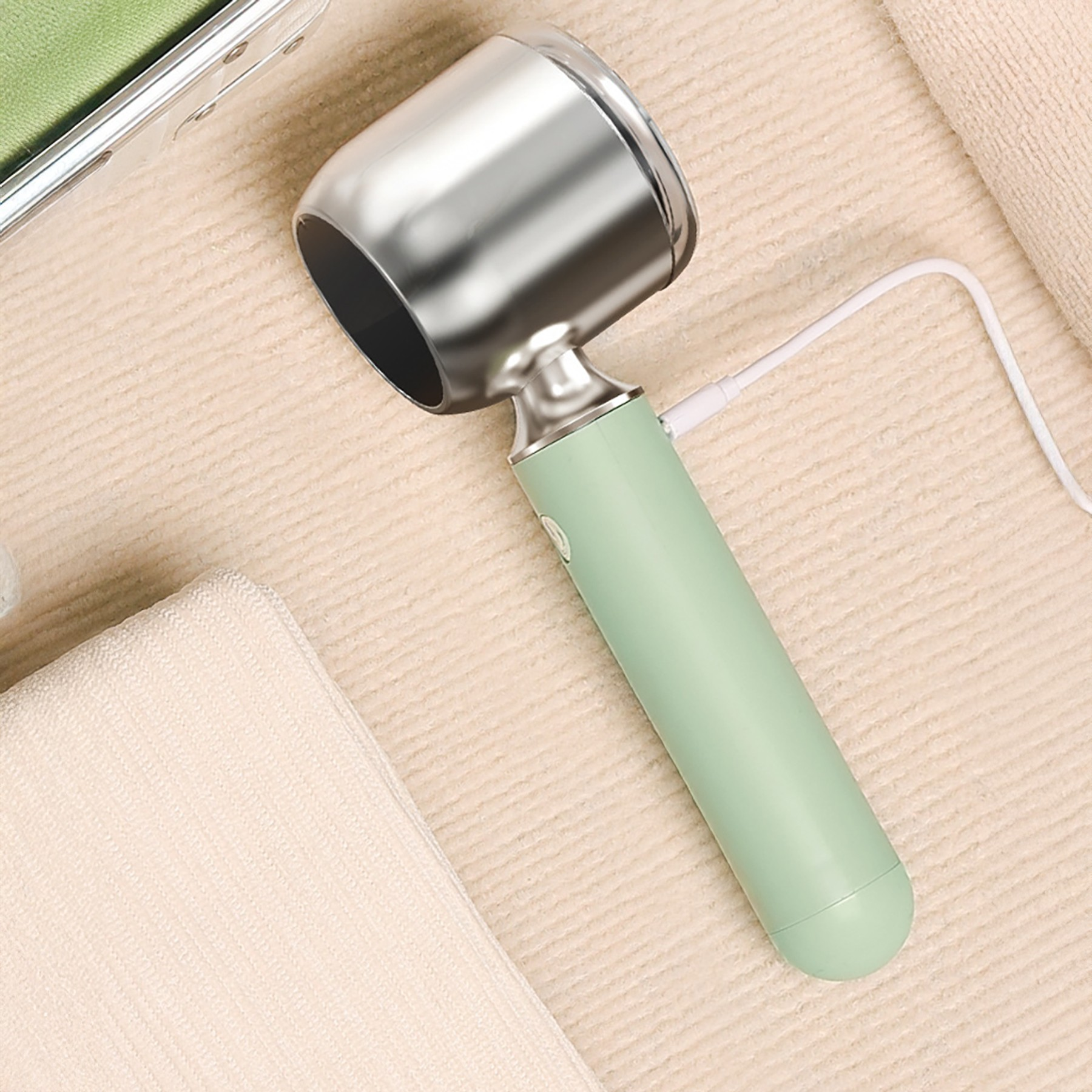USB şarj kablosuna sahip gümüş ve yeşil elektrikli tıraş makinesi bej bir havlu üzerinde duruyor.