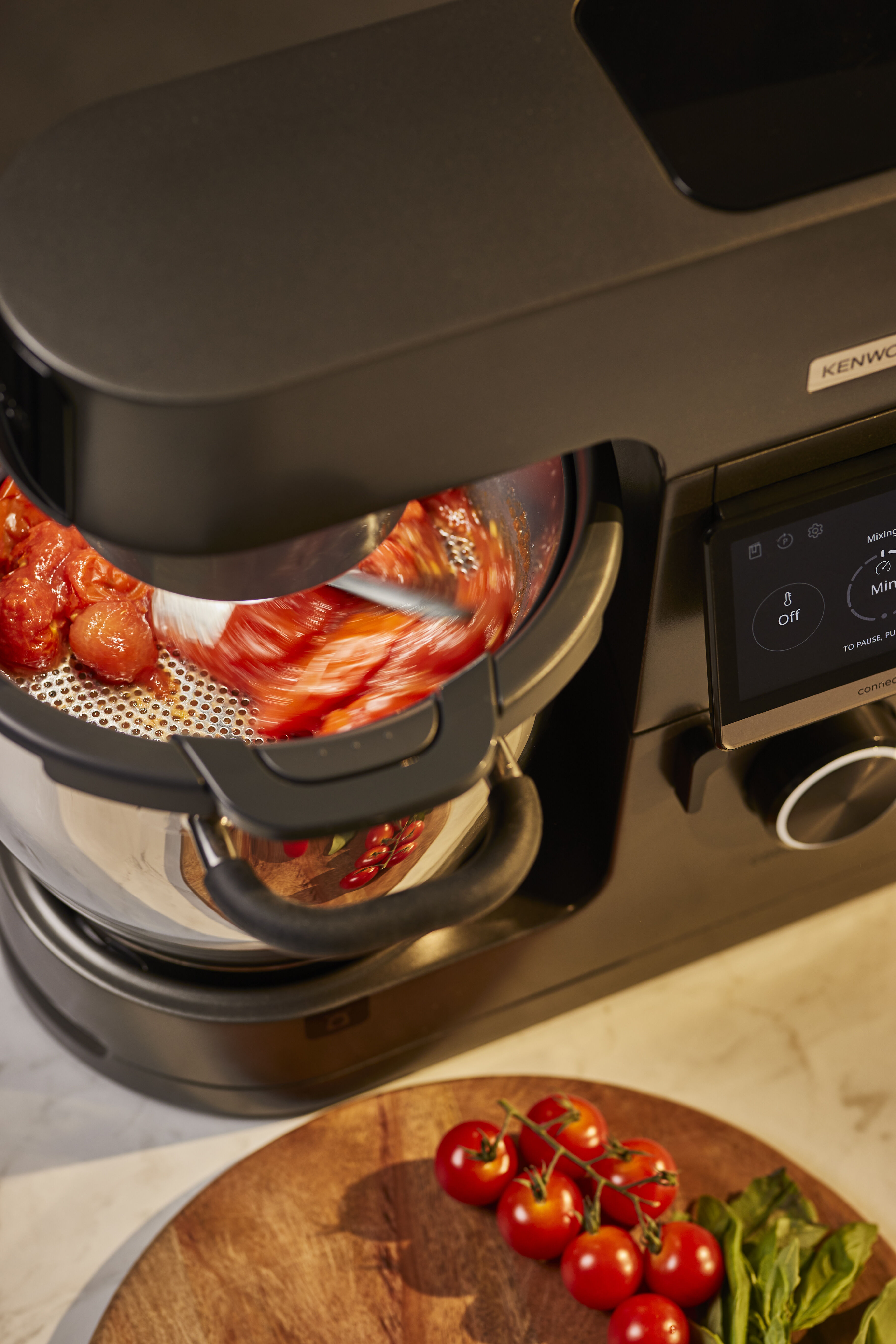 Eine Küchenmaschine verarbeitet Tomaten. Daneben liegen Kirschtomaten auf einem Holzbrett.