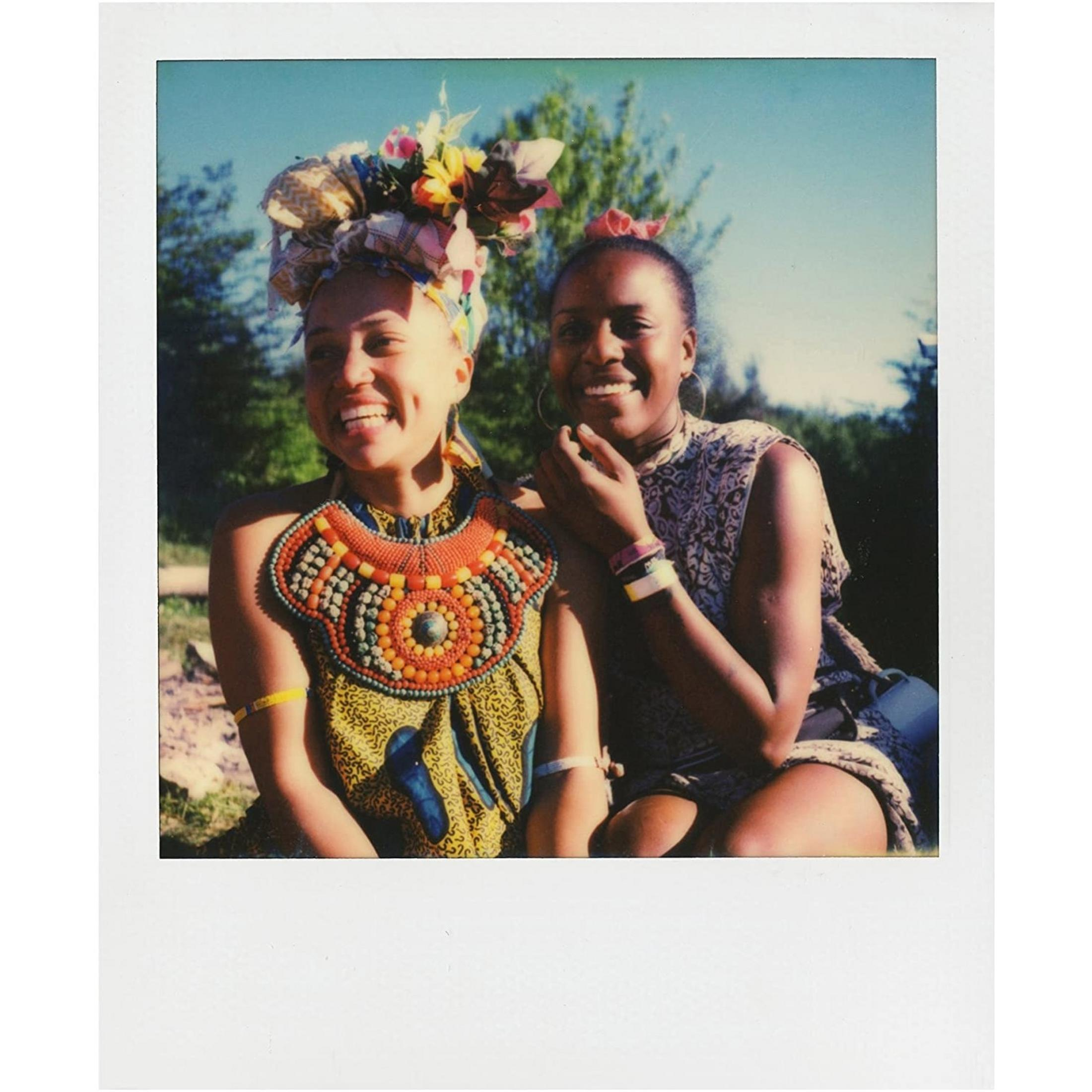 Dos mujeres sonriendo al aire libre, una con tocado floral y collar colorido.