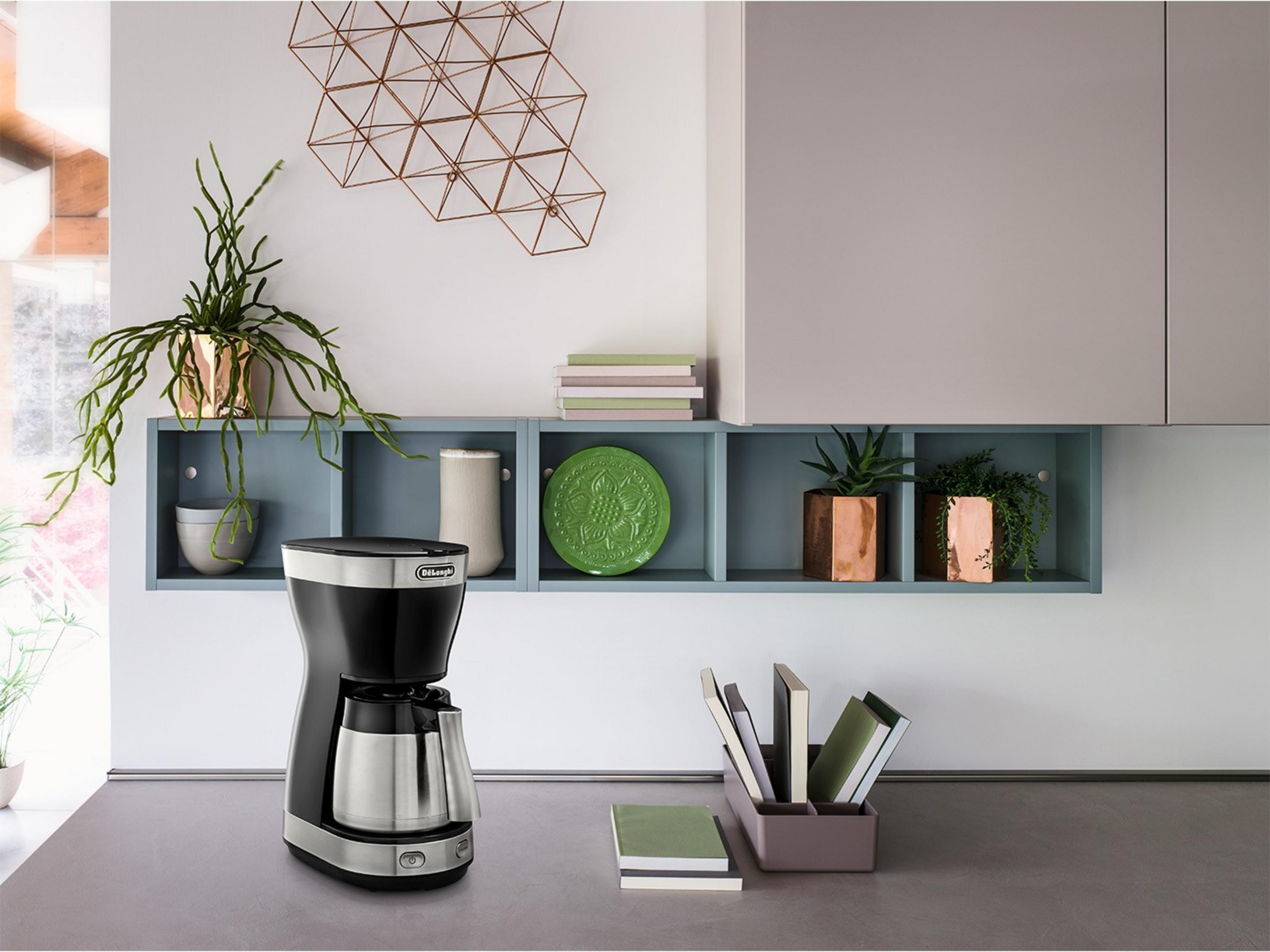 Ekspres do kawy stoi na szarym blacie obok książek i roślin.