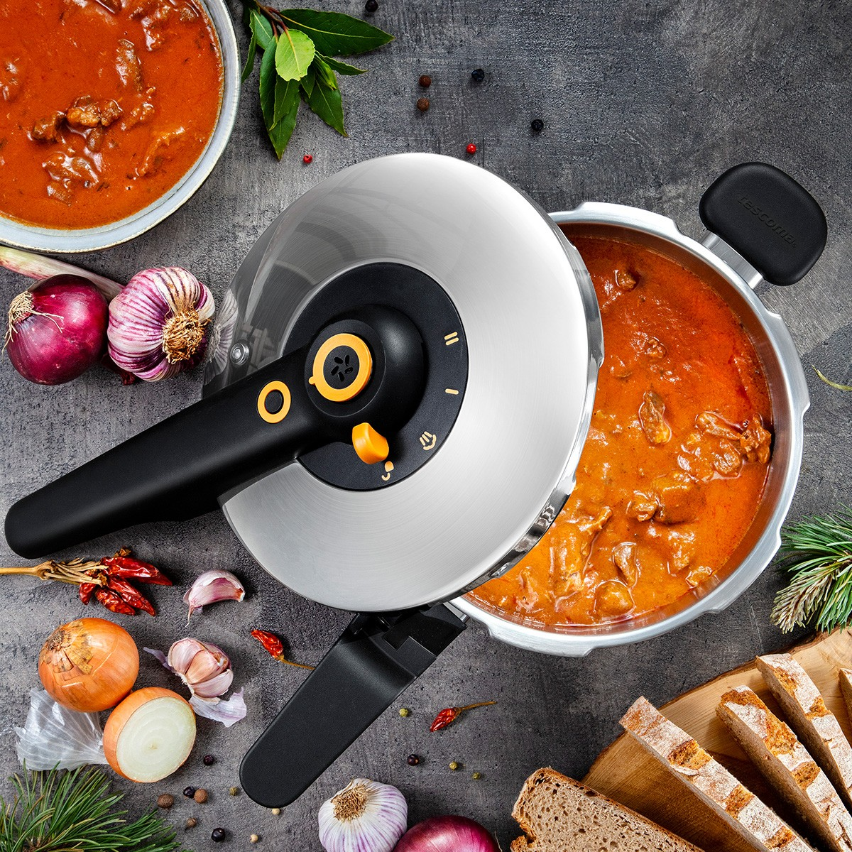 Ein Schnellkochtopf gefüllt mit Gulasch, neben einer Schüssel Gulasch, Zwiebeln, Knoblauch, Brot und Gewürzen.