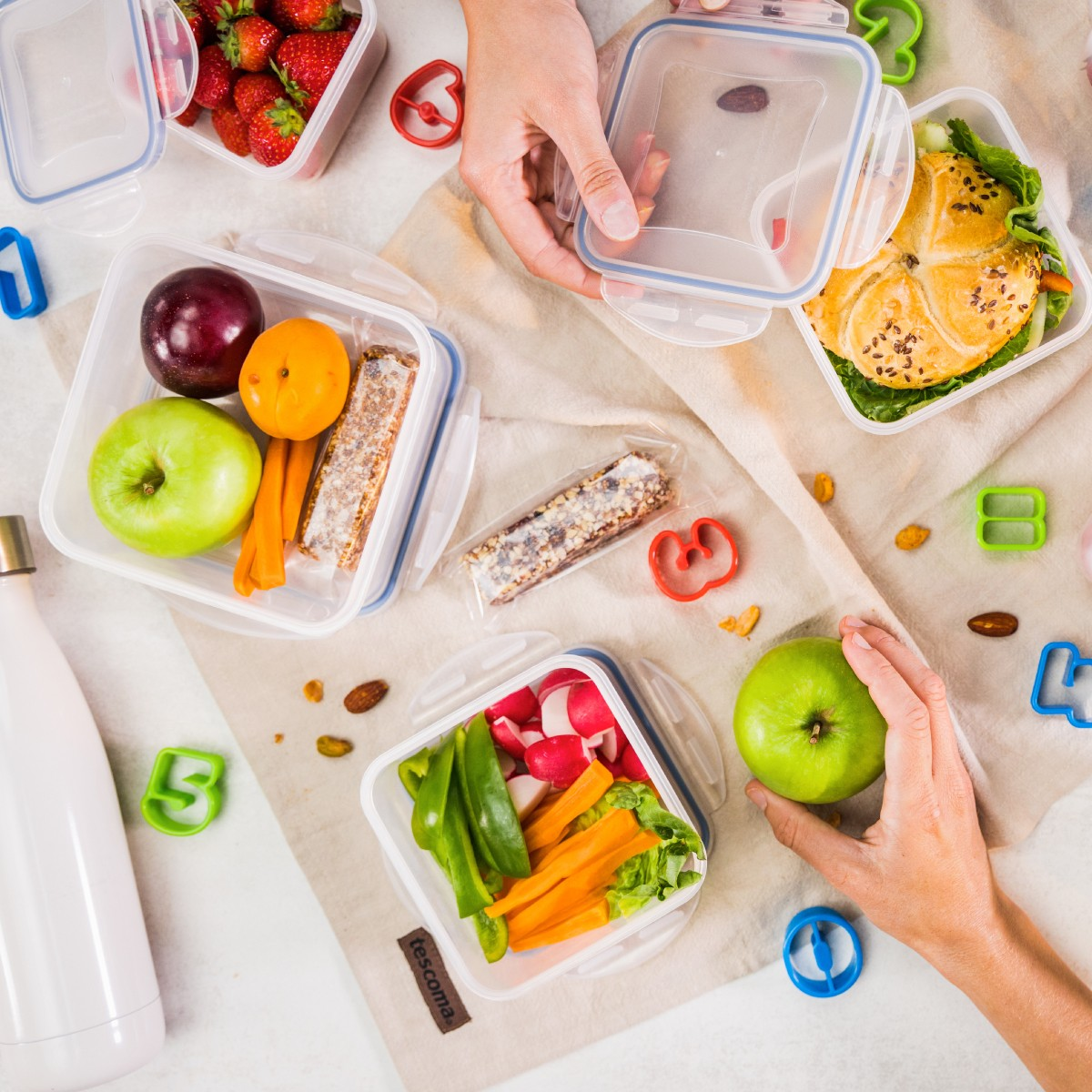 Bunte Brotdosen mit Obst, Gemüse, Sandwiches und Müsliriegeln, arrangiert auf einer Decke.
