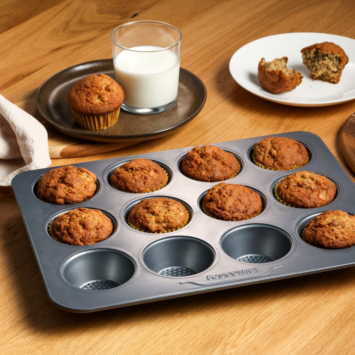 Ein Muffinblech mit acht Muffins, ein Glas Milch und zwei Muffins auf einem Teller.