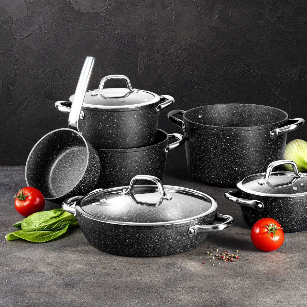 Granit-Kochgeschirr-Set mit Tomaten und Grünzeug auf dunklem Hintergrund.