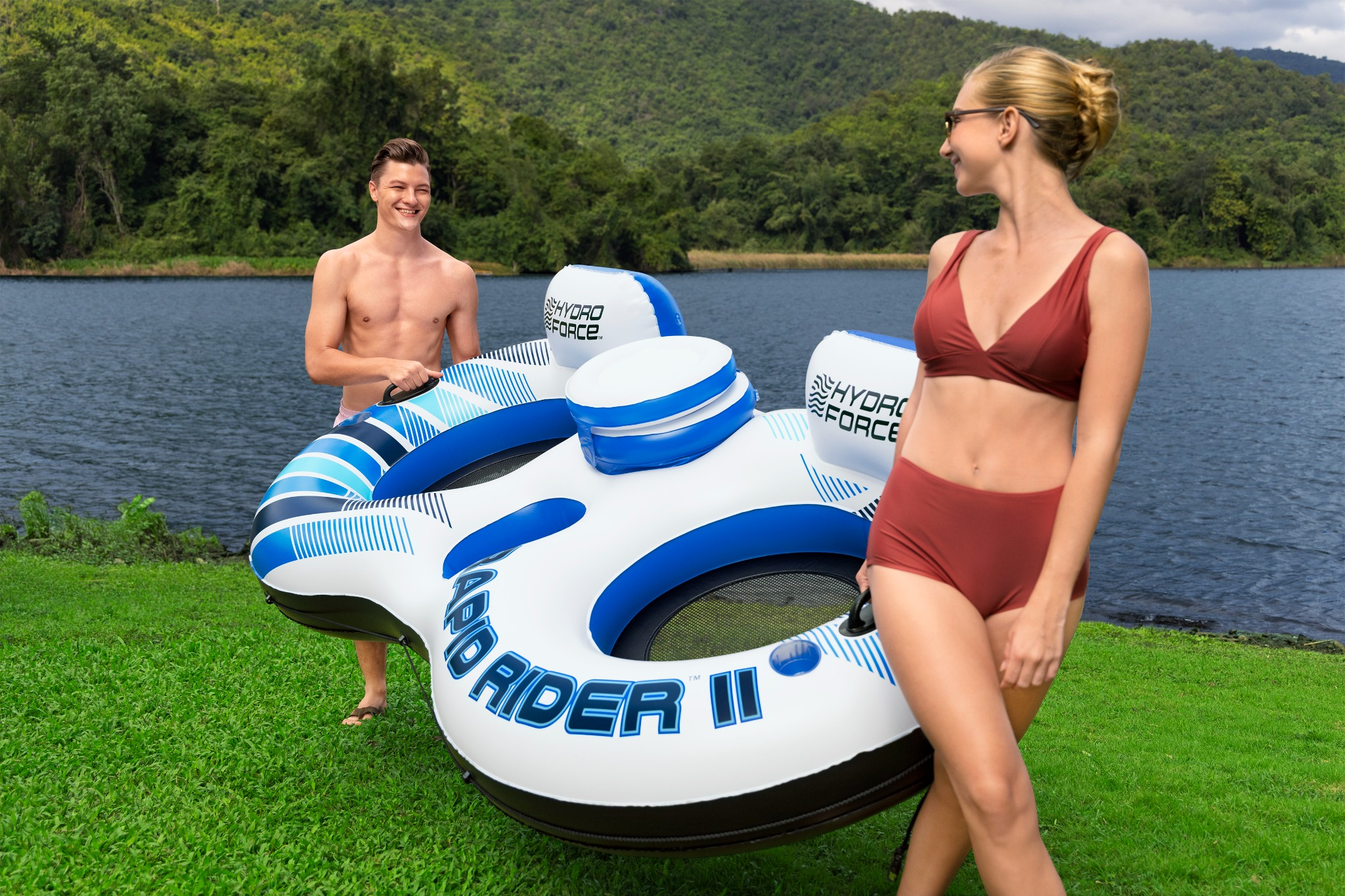 Mann und Frau halten ein blau-weißes Schlauchboot am Ufer eines Sees mit bewaldeten Hügeln im Hintergrund.