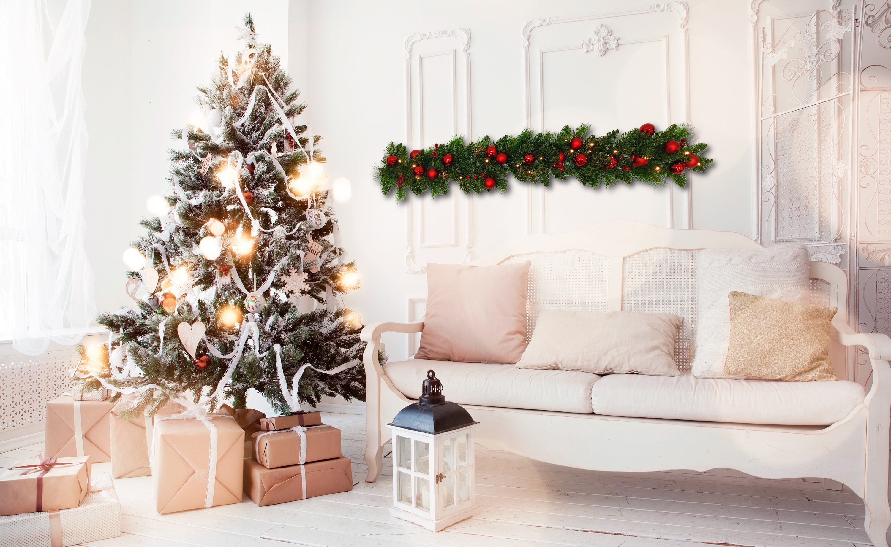 Ein geschmückter Weihnachtsbaum mit Lichtern und Geschenken neben einer weißen Couch mit Kissen.