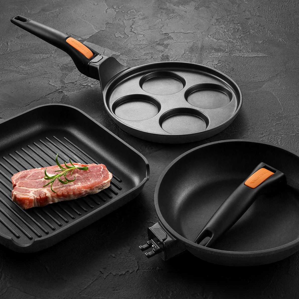 Drei schwarze Bratpfannen auf dunklem Untergrund. Eine mit Fleisch und Rosmarin.