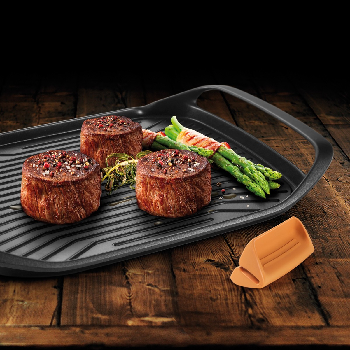 Drei saftige Steaks mit Gewürzen auf einem Grill, daneben Spargel und Kräuter.