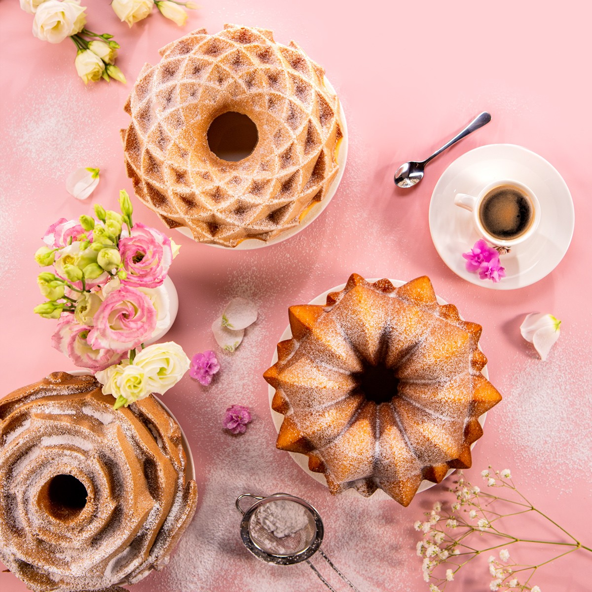 Drei Gugelhupfe mit Puderzucker, Blumen und Kaffee auf rosa Hintergrund.