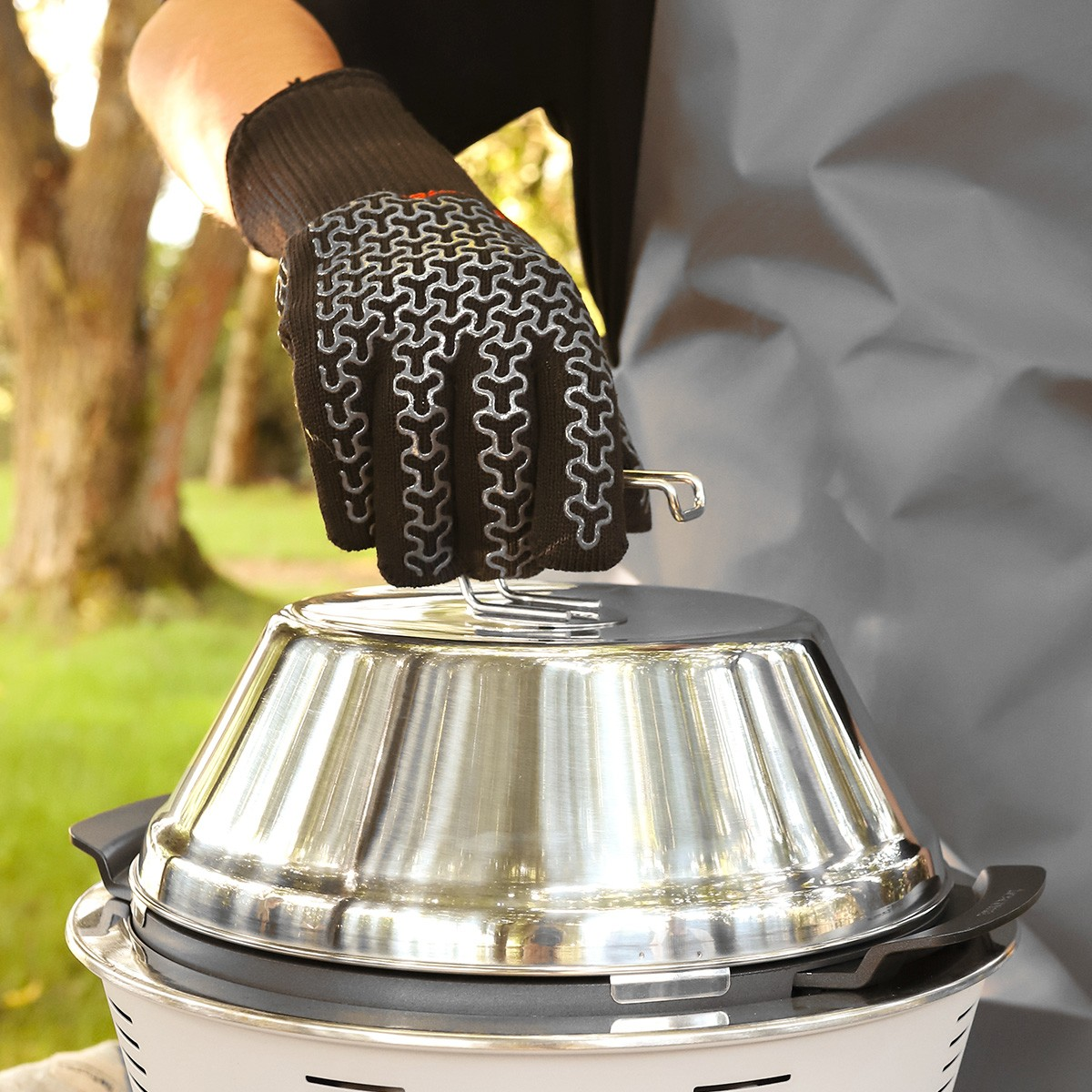 Eine Hand in einem Grillhandschuh hebt den Edelstahldeckel eines Grills an.