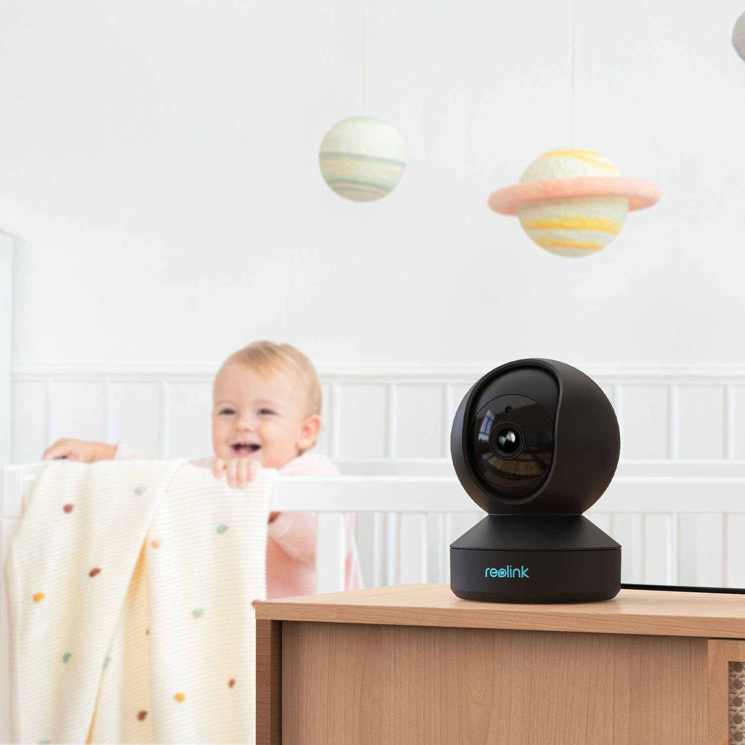 Eine schwarze Reolink-Kamera steht auf einem Holztisch vor einem Baby im Kinderbett.