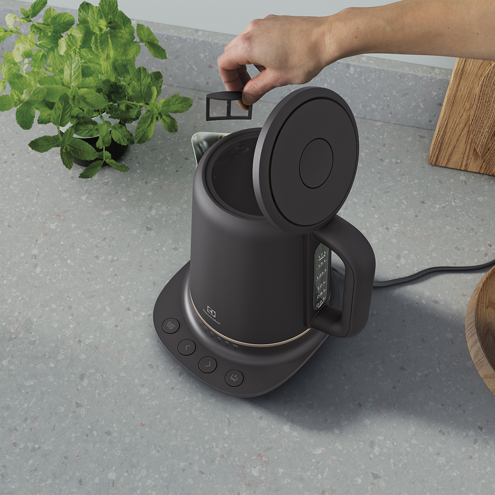 Ręka wkłada czarny filtr do herbaty do czajnika. W pobliżu roślina mięty.