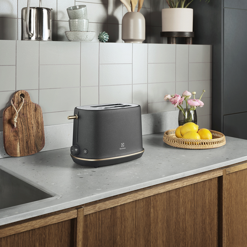 Nowoczesny, ciemny toster na kamiennym blacie obok cytryn i kwiatów.