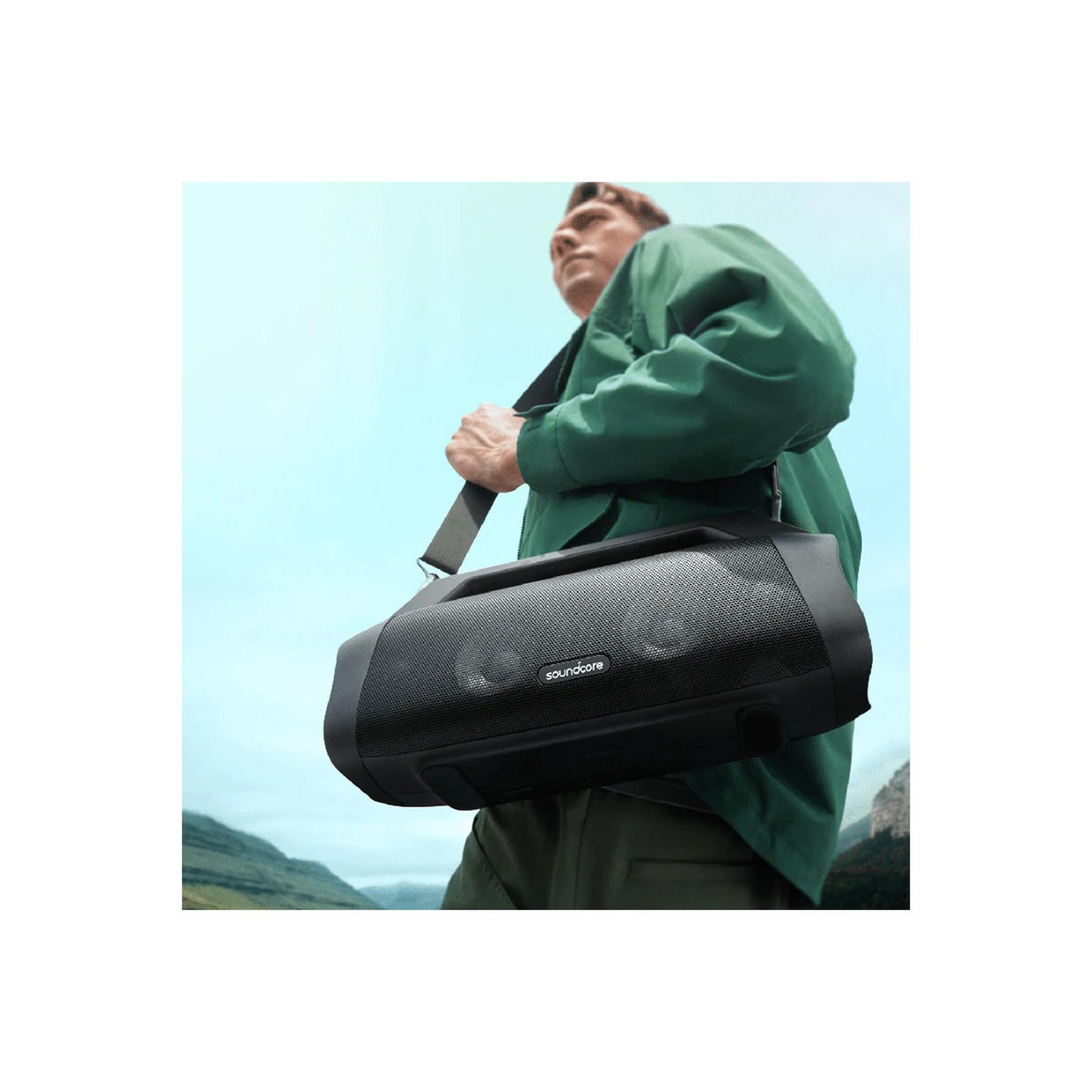 Uomo con giacca verde porta altoparlante nero a spalla, montagne sullo sfondo.