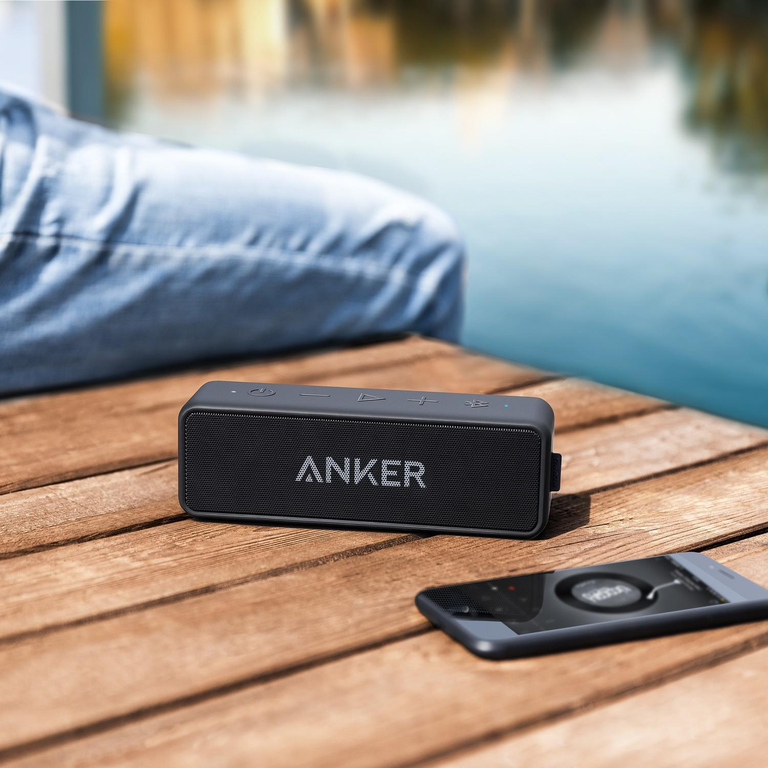 Un altoparlante Anker nero e uno smartphone poggiano su un molo di legno vicino all'acqua.