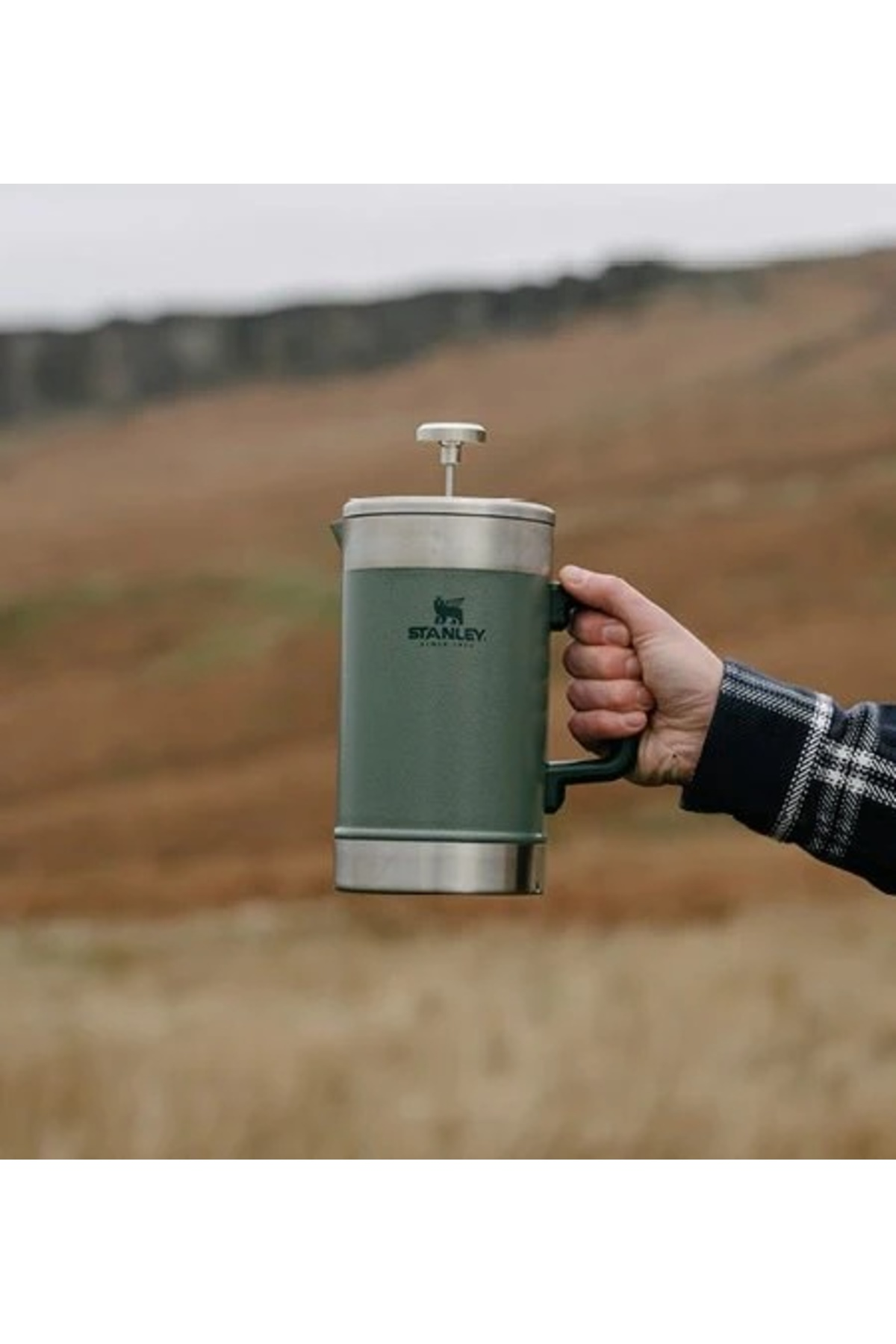 Una French press Stanley verde è tenuta in mano, con paesaggio sullo sfondo.