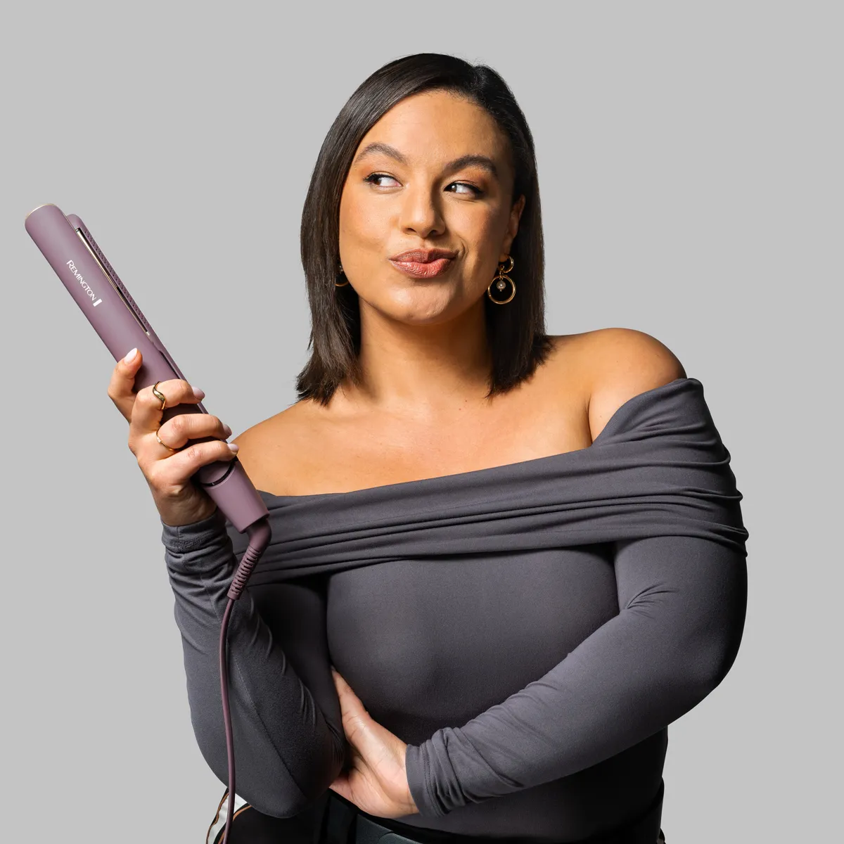 Una donna tiene una piastra per capelli viola, indossa un top grigio senza spalline.