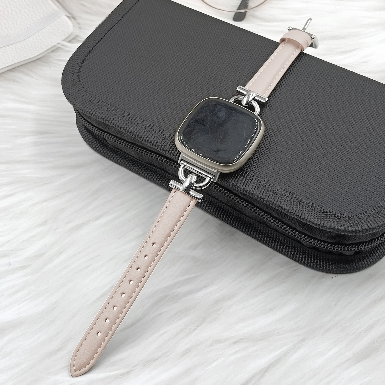 Eine rechteckige Smartwatch mit hellem Band liegt auf einer schwarzen Tasche.