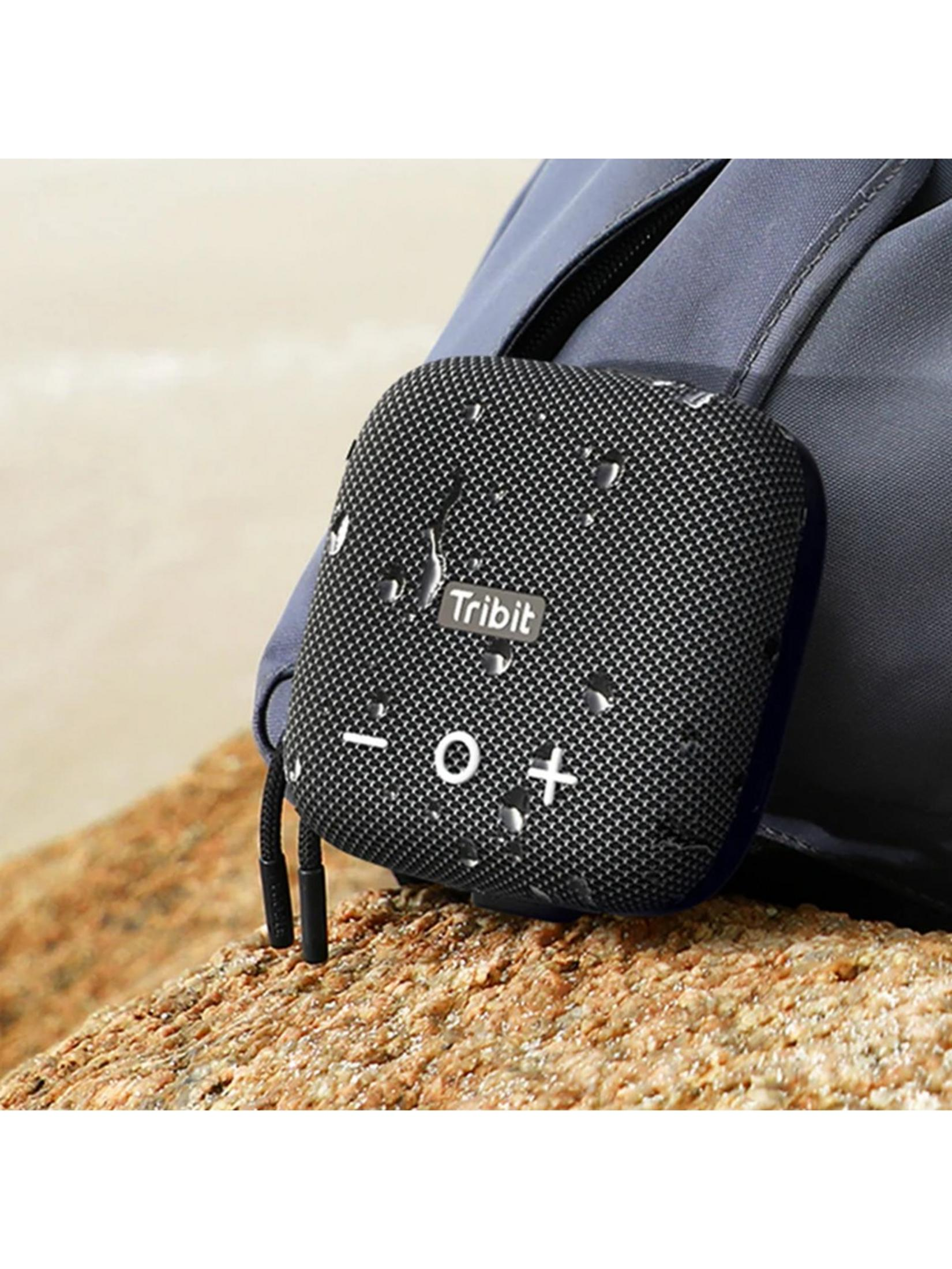 Ein schwarzer Tribit-Lautsprecher mit Wassertropfen sitzt auf einem Stein neben einem Rucksack.