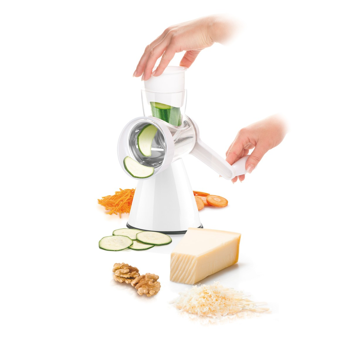 Eine weiße Reibe verarbeitet eine Gurke zu Scheiben. Daneben liegen Karotten, Nüsse und Käse.