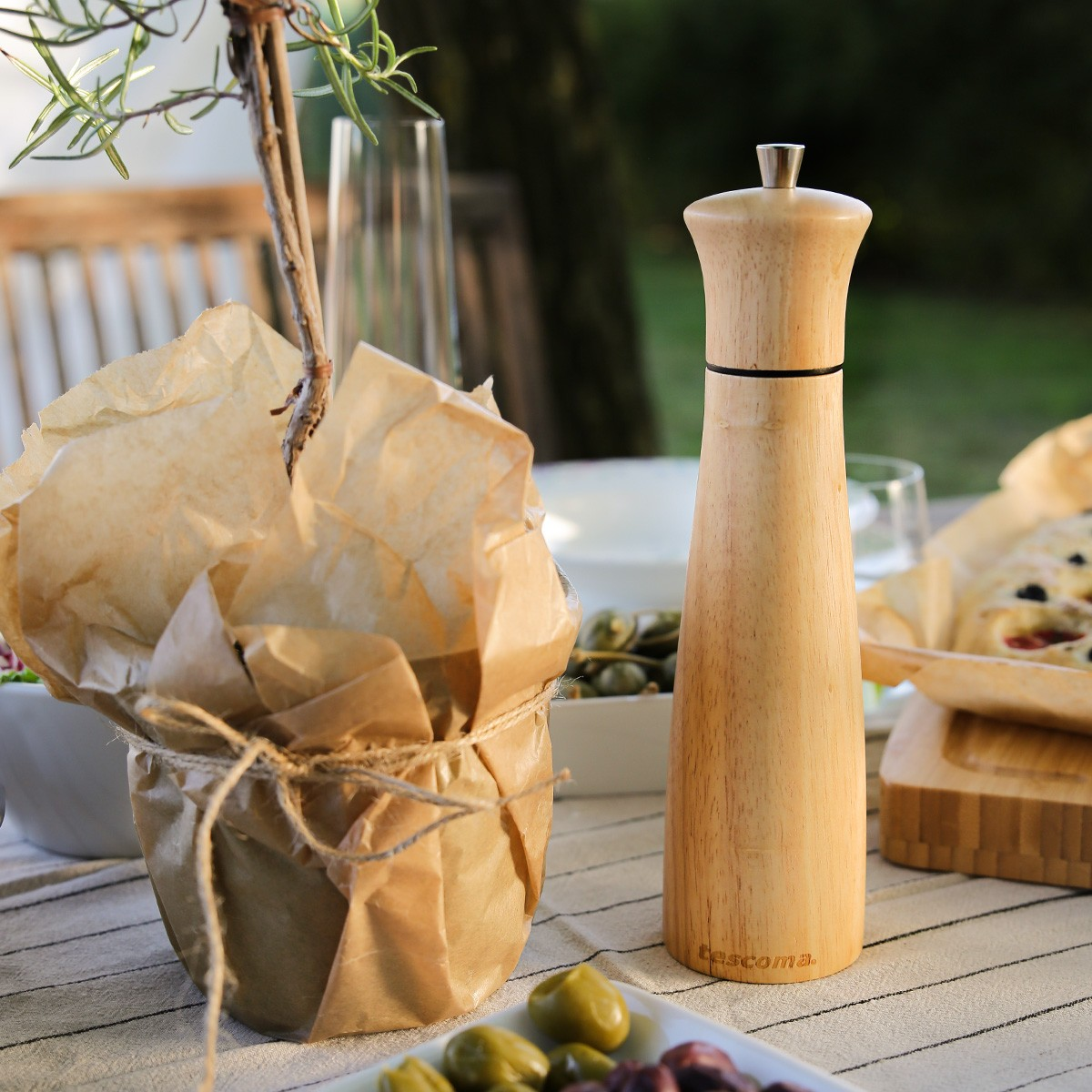 Eine Holzpfeffermühle steht auf einem Tisch mit Oliven, Brot und einer Topfpflanze.