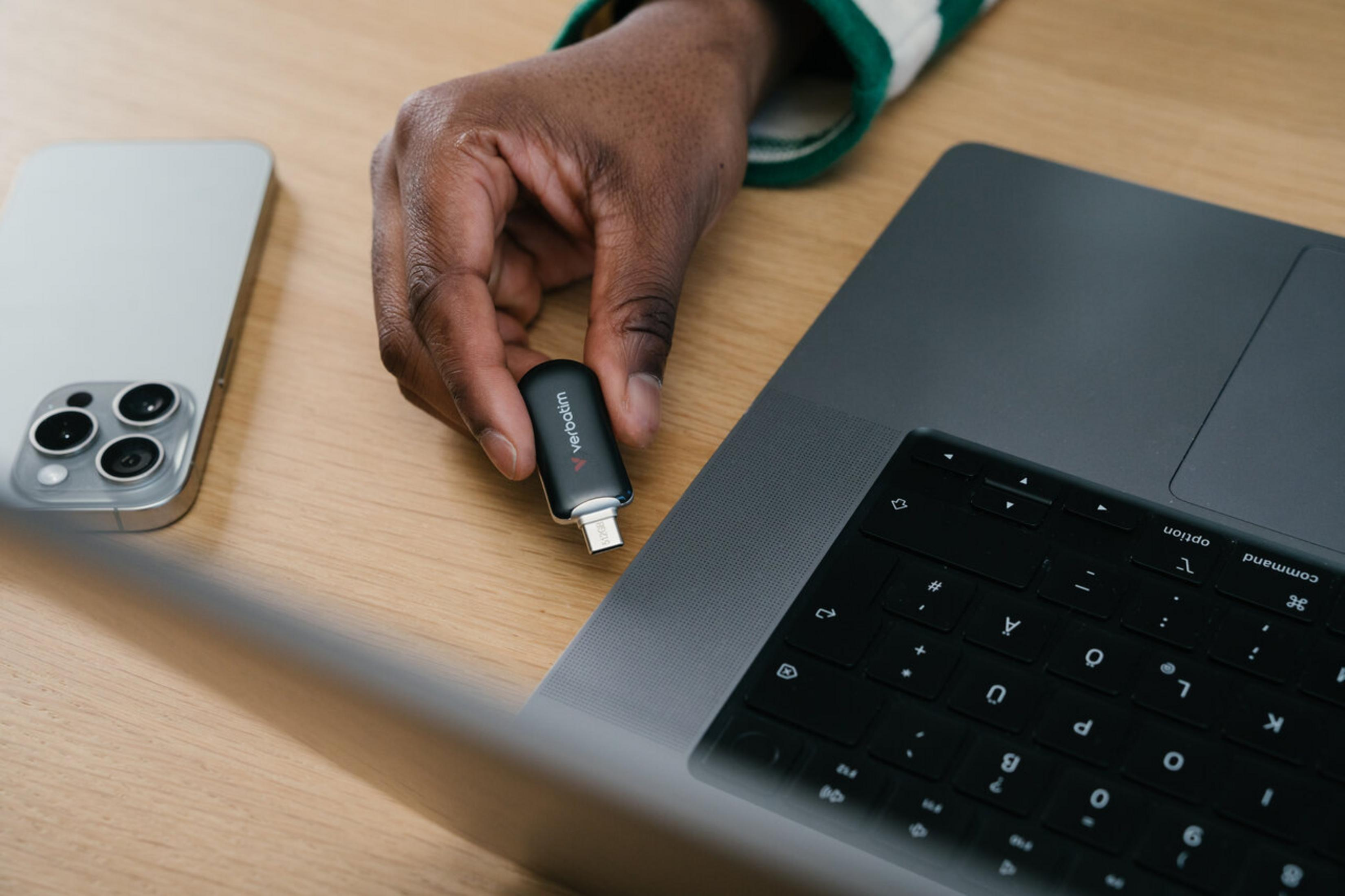 Dłoń trzyma pendrive Verbatim obok laptopa i iPhone'a.