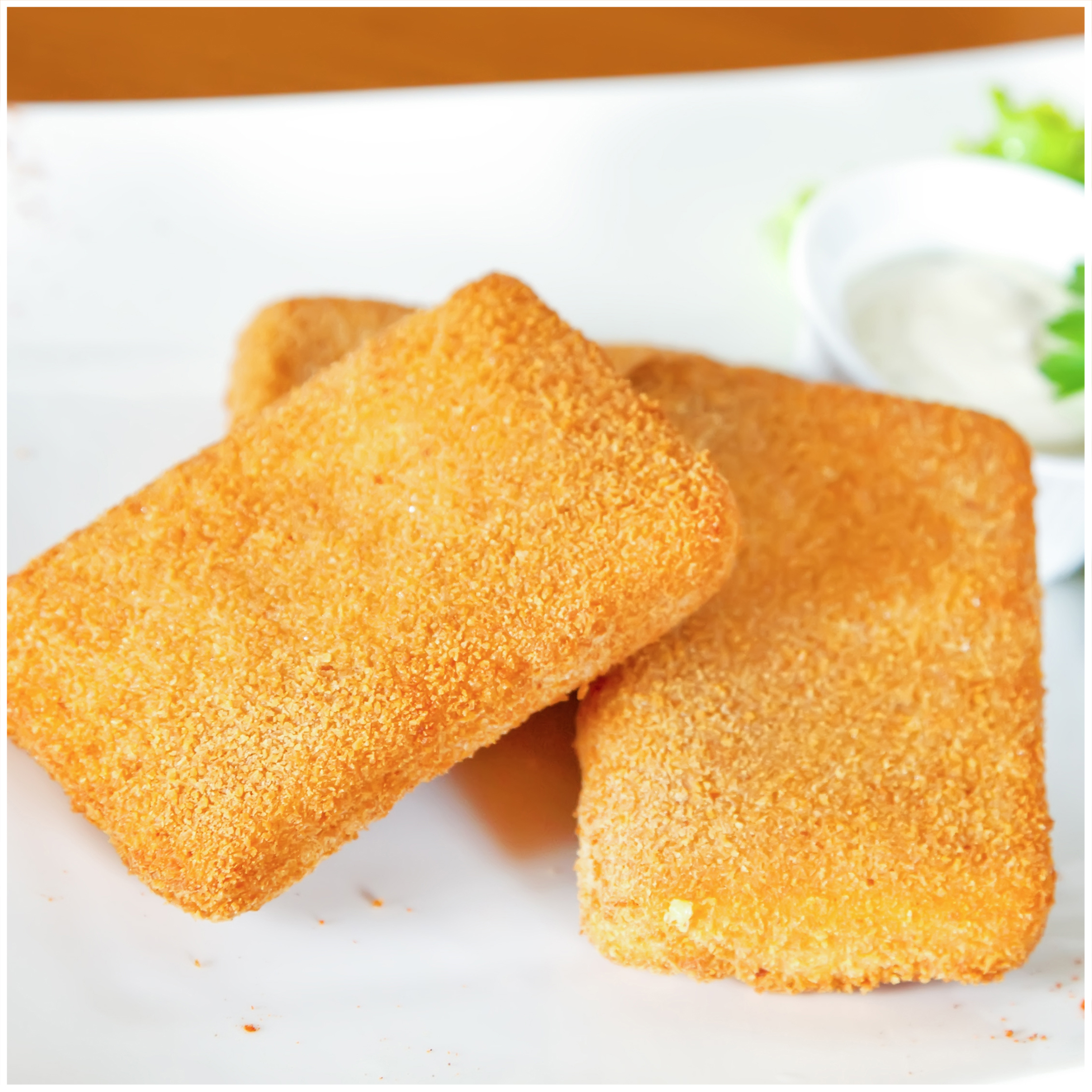 Drie goudbruine gefrituurde kaaskubus op een wit bord met saus en sla.
