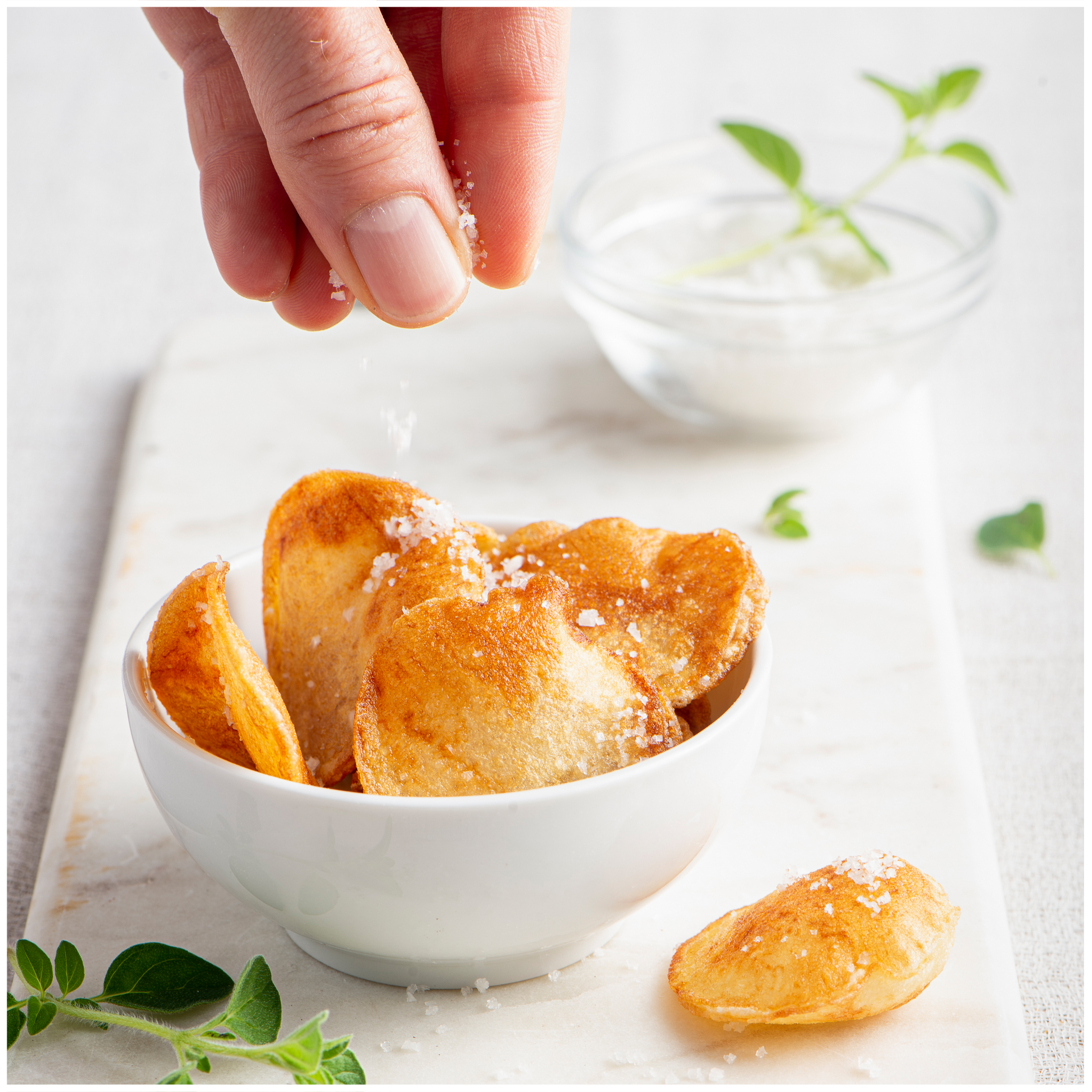 Salt is being sprinkled onto crispy potato chips in a white bowl.