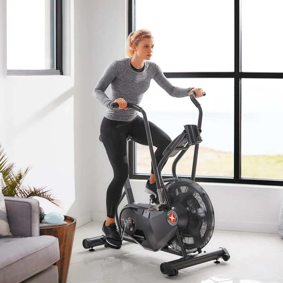 Une femme s'entraîne sur un vélo d'appartement à entraînement par ventilateur dans une pièce lumineuse.